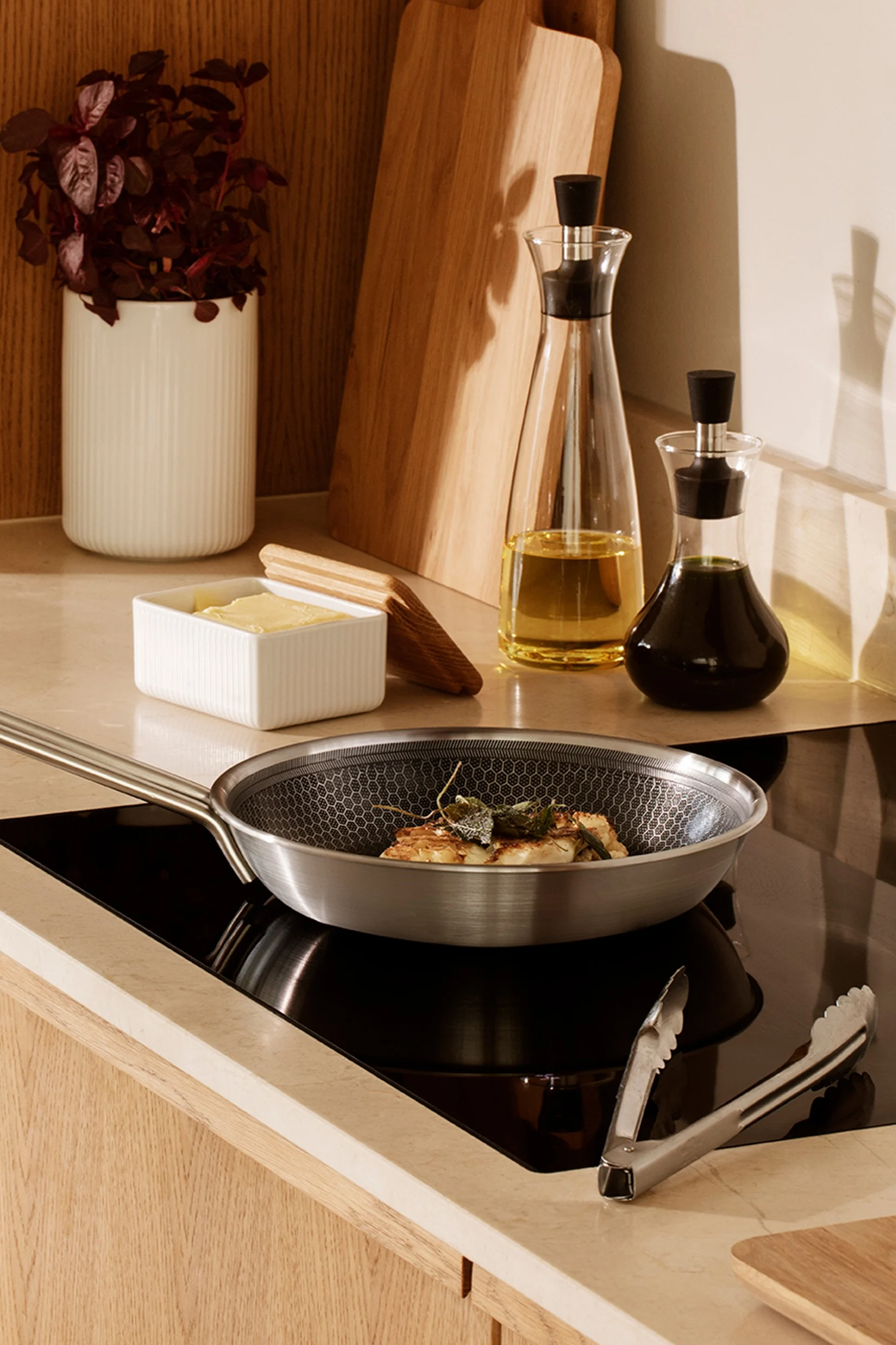 Stainless steel frying pan with food on an induction hob, alongside butter, oil bottles, and kitchen tools in a modern kitchen.