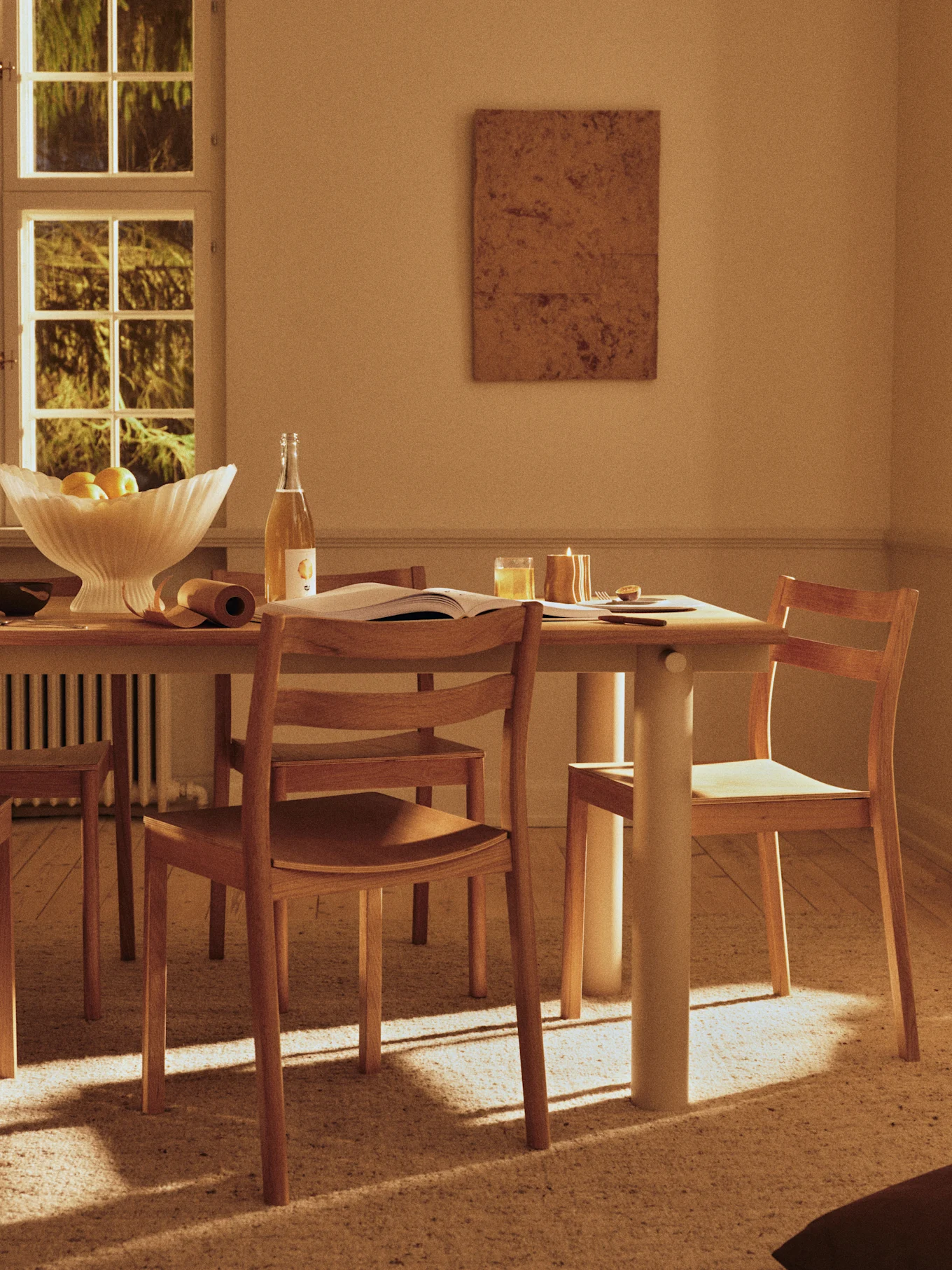 A sunlit dining room with a wooden table, chairs, a glass bowl of fruit, a drink bottle, and a lit candle on the table, casting long shadows on the floor.