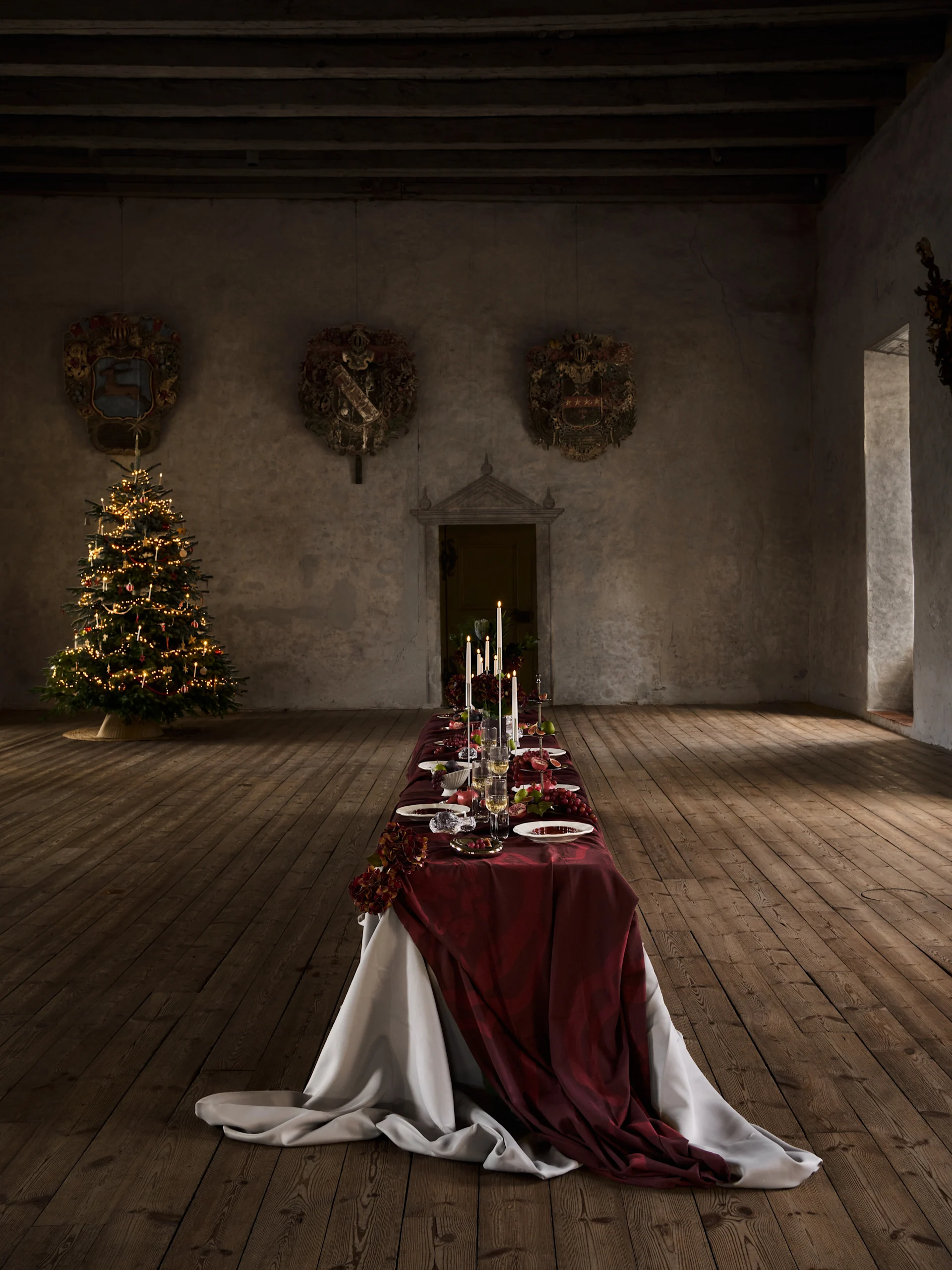 Long Christmas dinner table with red cloth, candles, fruit, and settings, next to a lighted Christmas tree in an old hall.