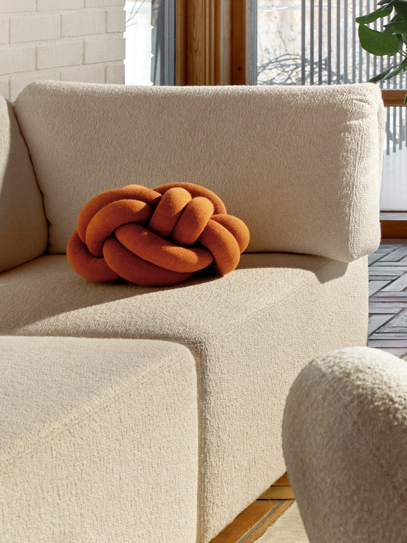 An orange knot pillow on a beige textured sofa, with a white brick wall and a window in the background.
