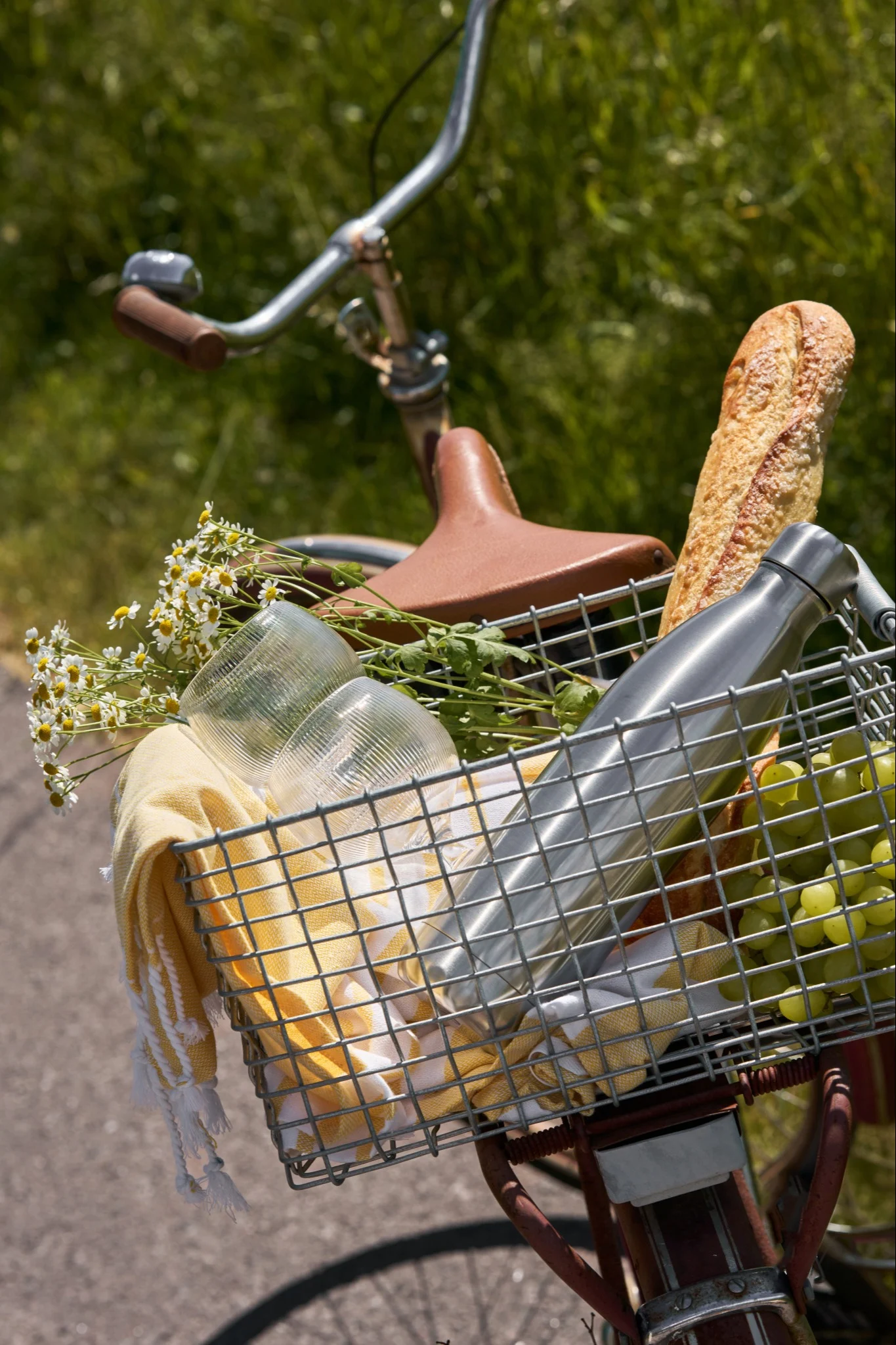 Bicycle with a front basket filled with picnic items: bread, green grapes, a silver water bottle, cups, a yellow towel, and flowers.