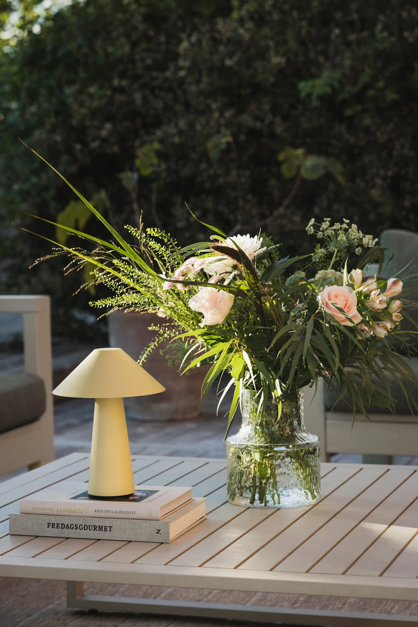 A yellow mushroom lamp, two stacked books, and a glass vase with pink roses and white flowers on a slatted outdoor table.