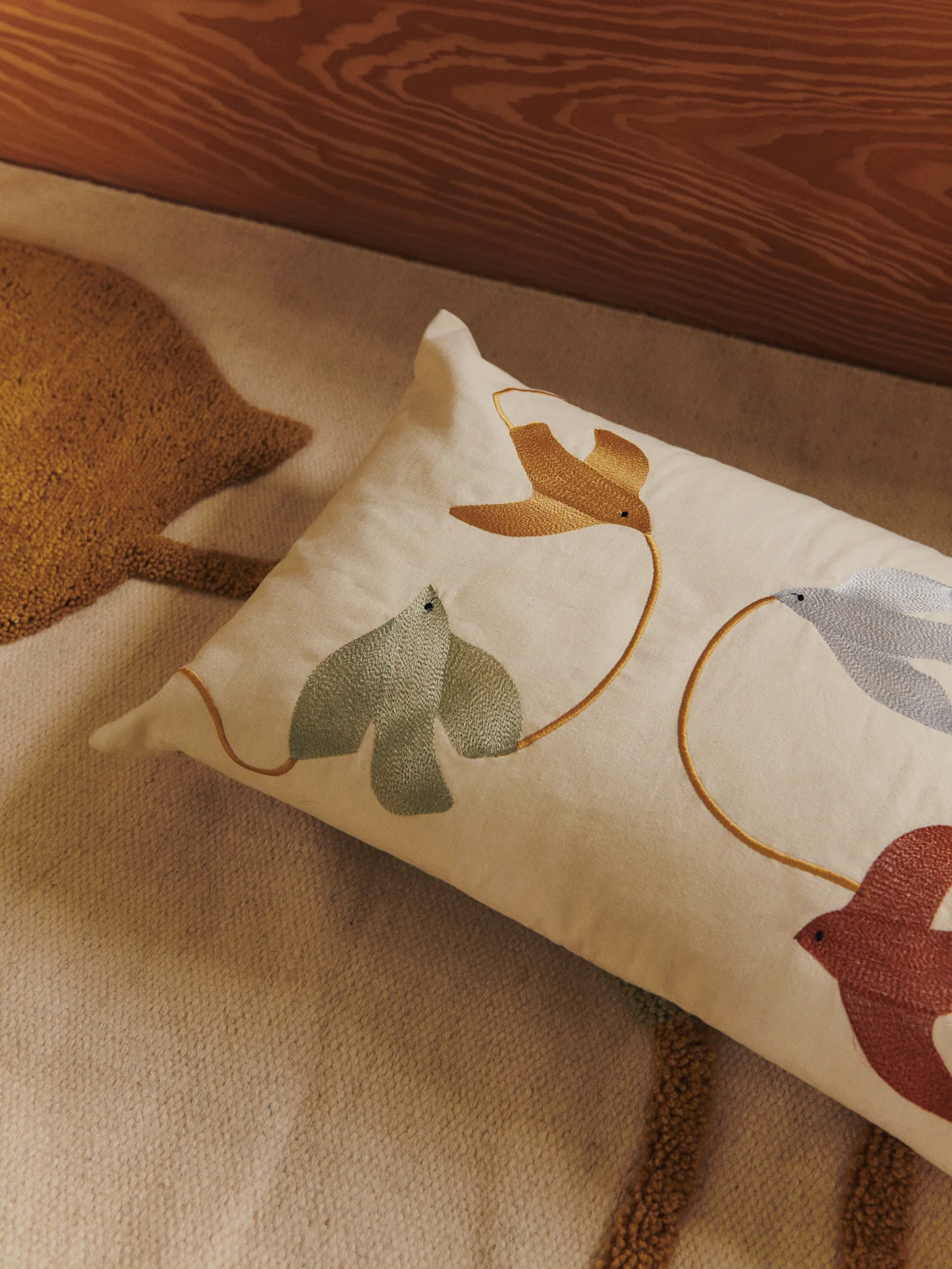 Lumbar pillow with embroidered birds and leaves in gold, green, blue, and reddish-brown on a light-colored rug.