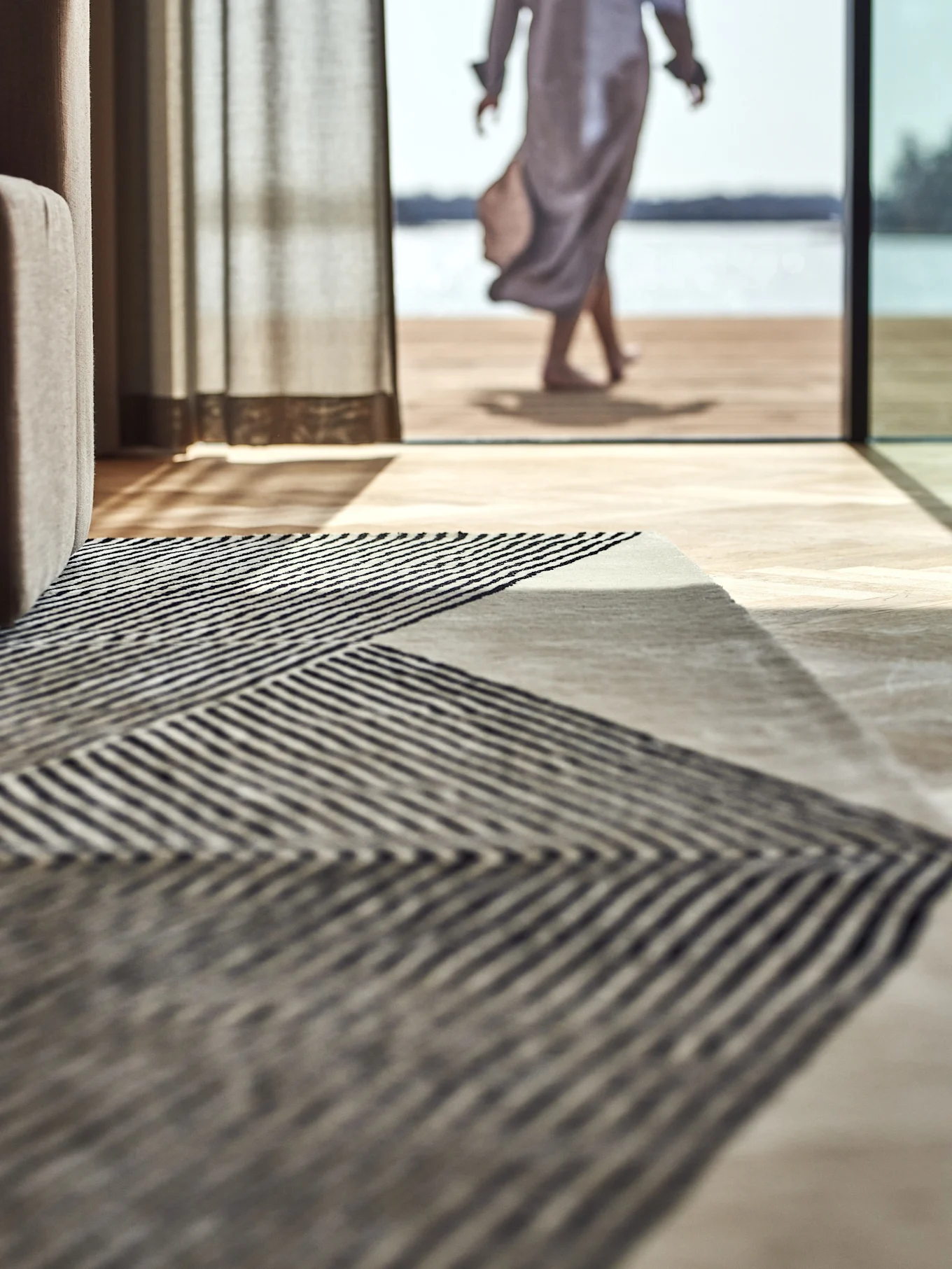 Geometric striped rug on a wooden floor, with a person walking towards a lake outside.