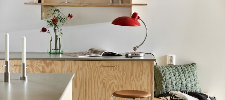 Here you see the Kaiser table lamp in ruby red with the Ikeru vase  in brushed steel, both from Fritz Hansen, in a modern plywood kitchen. 