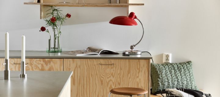 Here you see the Kaiser table lamp in ruby red with the Ikeru vase  in brushed steel, both from Fritz Hansen, in a modern plywood kitchen. 
