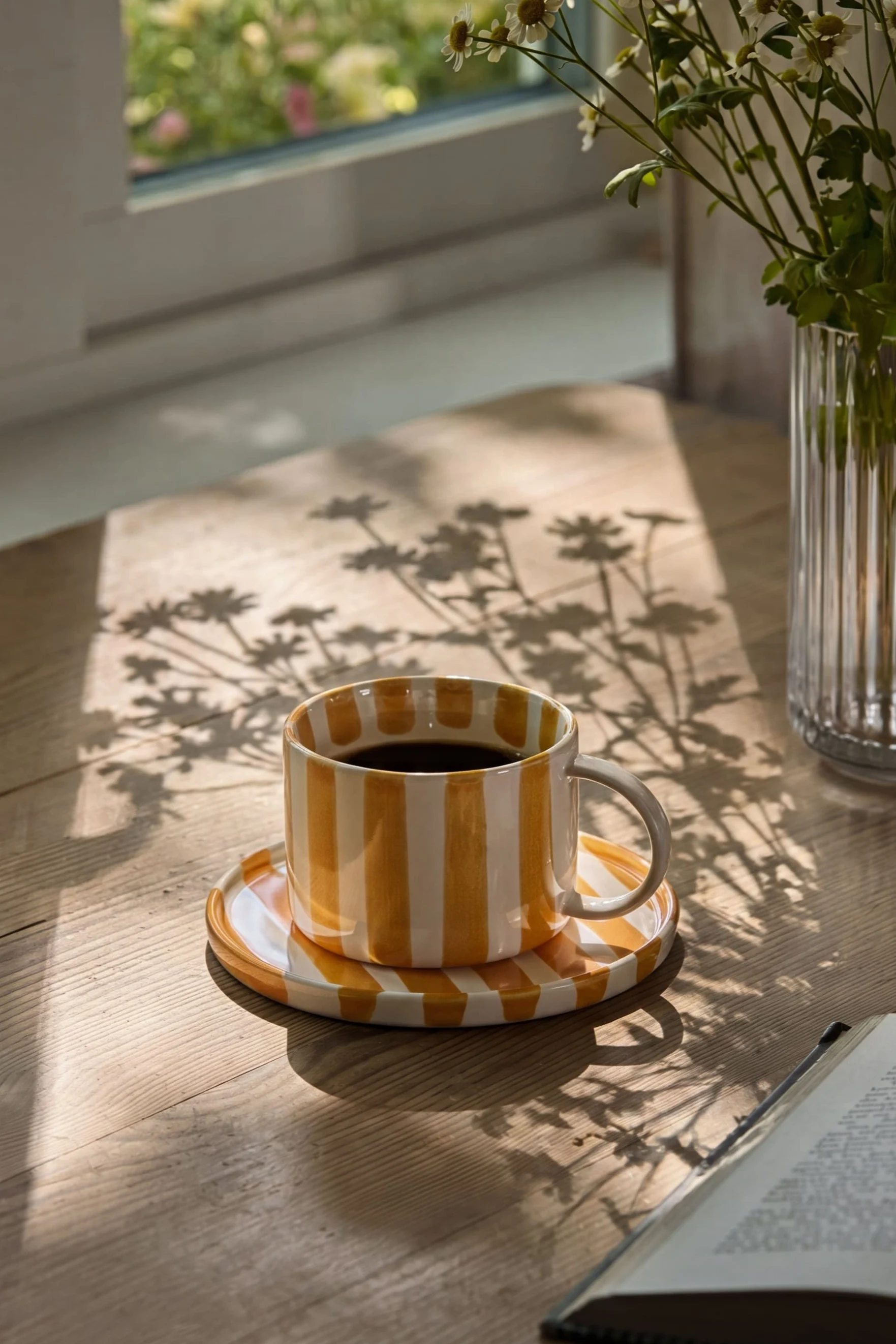 Yellow and white striped coffee mug on a wooden table with sun-dappled floral shadows, next to a vase of daisies and an open book.