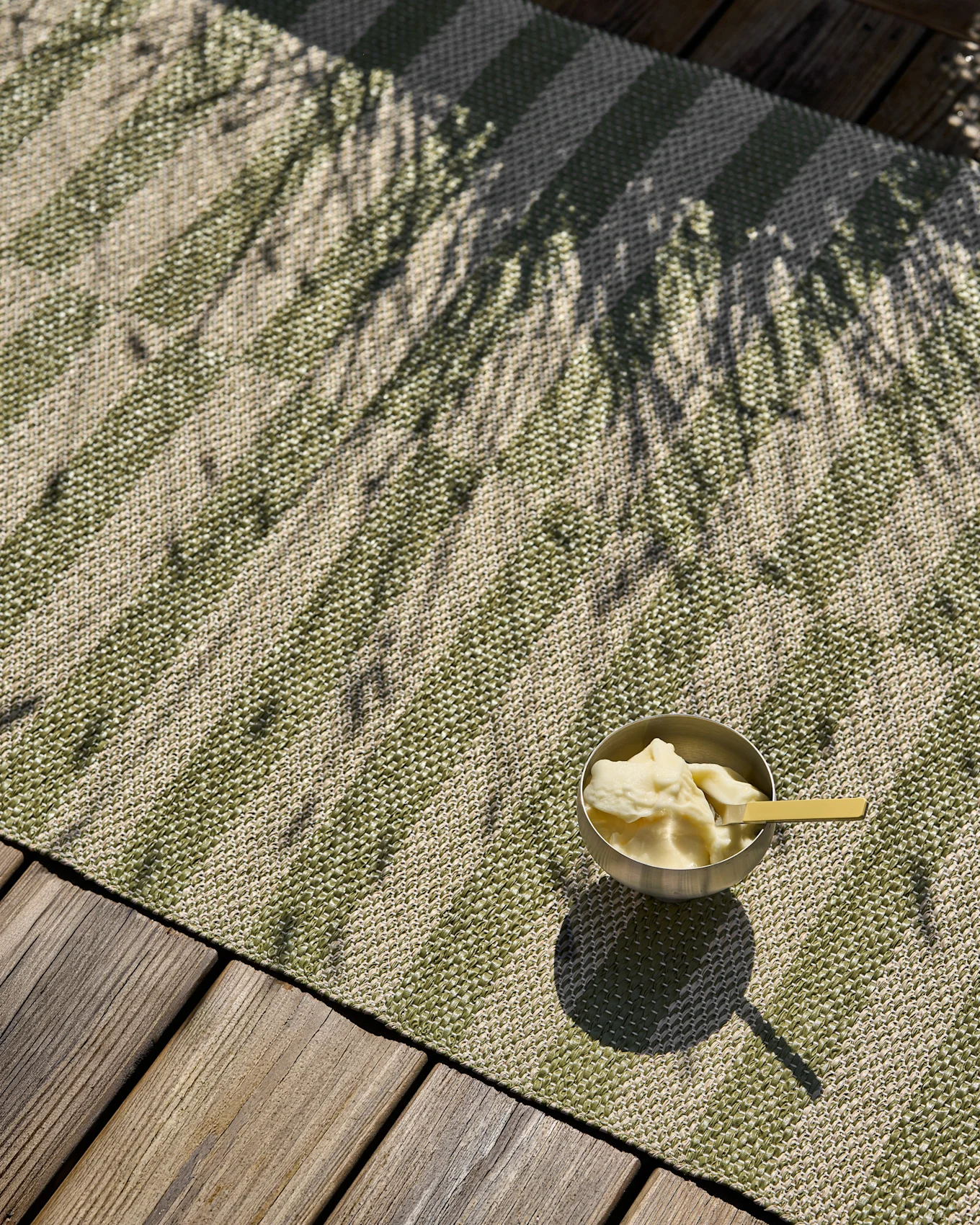 Green and beige striped woven rug with a bowl of ice cream and palm shadows on a wooden deck.