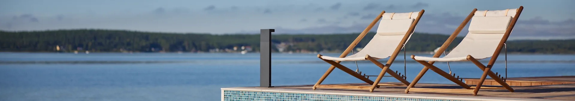 Two white fabric and wooden deck chairs on a deck overlooking a lake with a forest in the distance.