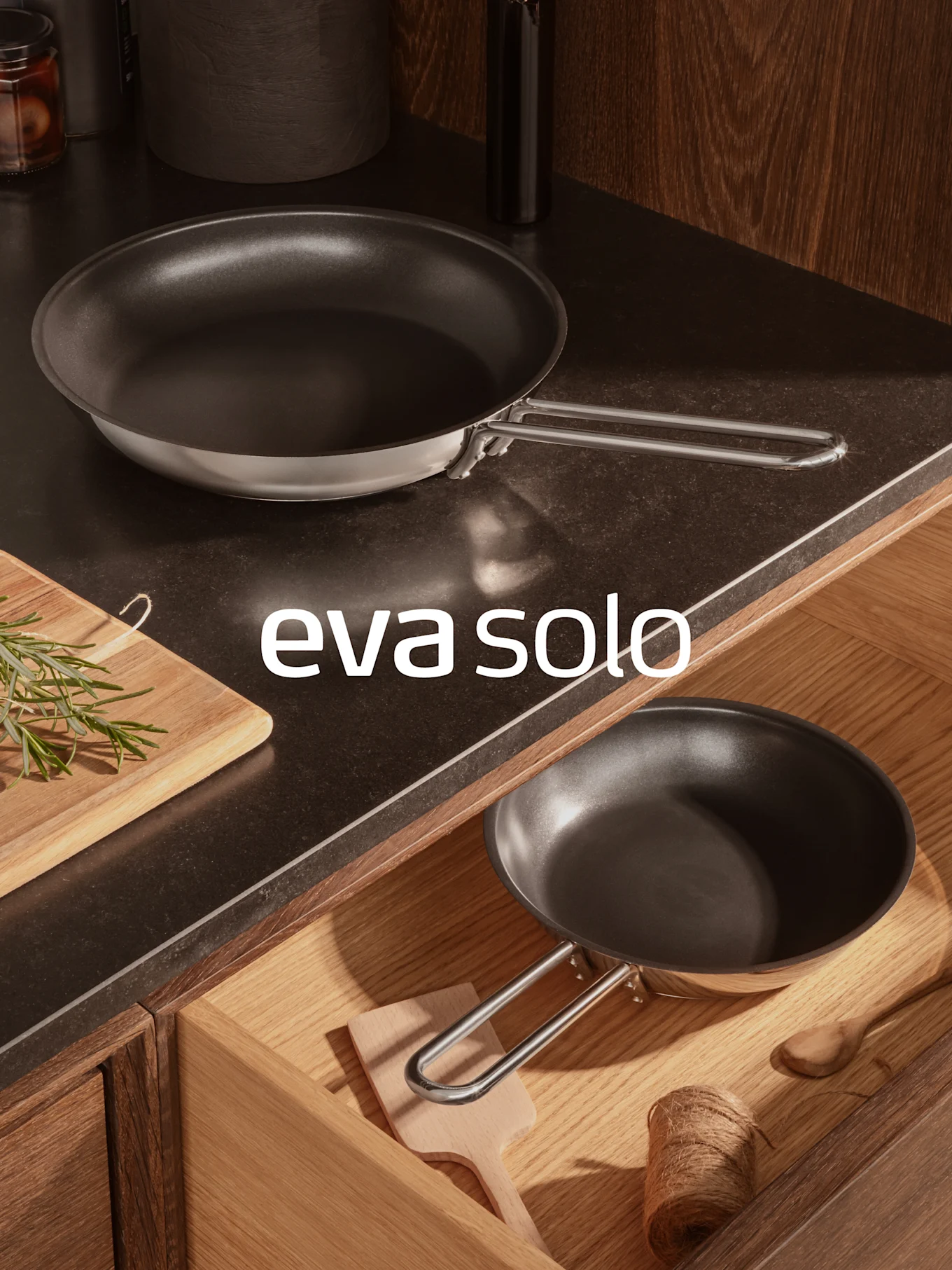 Two black frying pans, one on a dark kitchen counter with rosemary, one in an open wooden drawer. The 'eva solo' logo is visible.