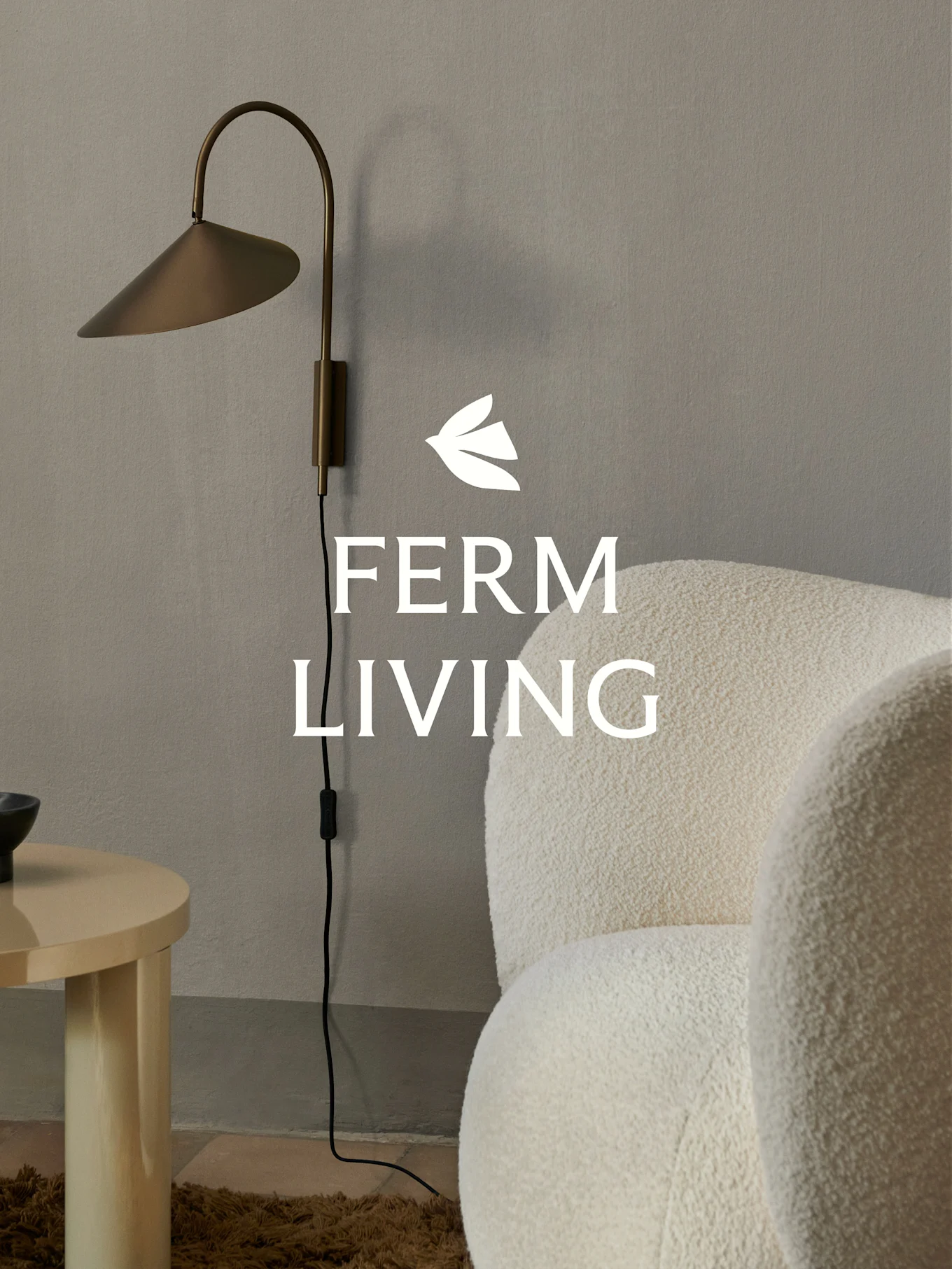 Ferm Living logo over a brass wall lamp, a cream boucle armchair, and a light wood side table in a minimalist setting.