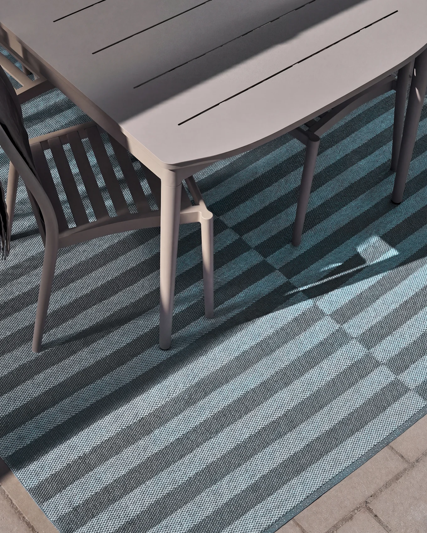 Overhead view of an outdoor dining table and chair on a striped rug with light and dark blue stripes, partially illuminated by sunlight.