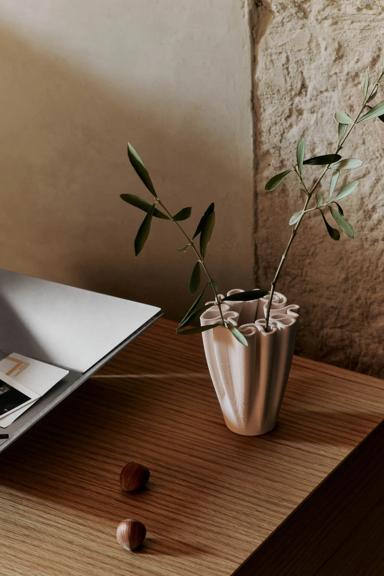 Light pink ruffled vase with green branches, two hazelnuts, and a grey tray on a wooden table next to a rustic wall.