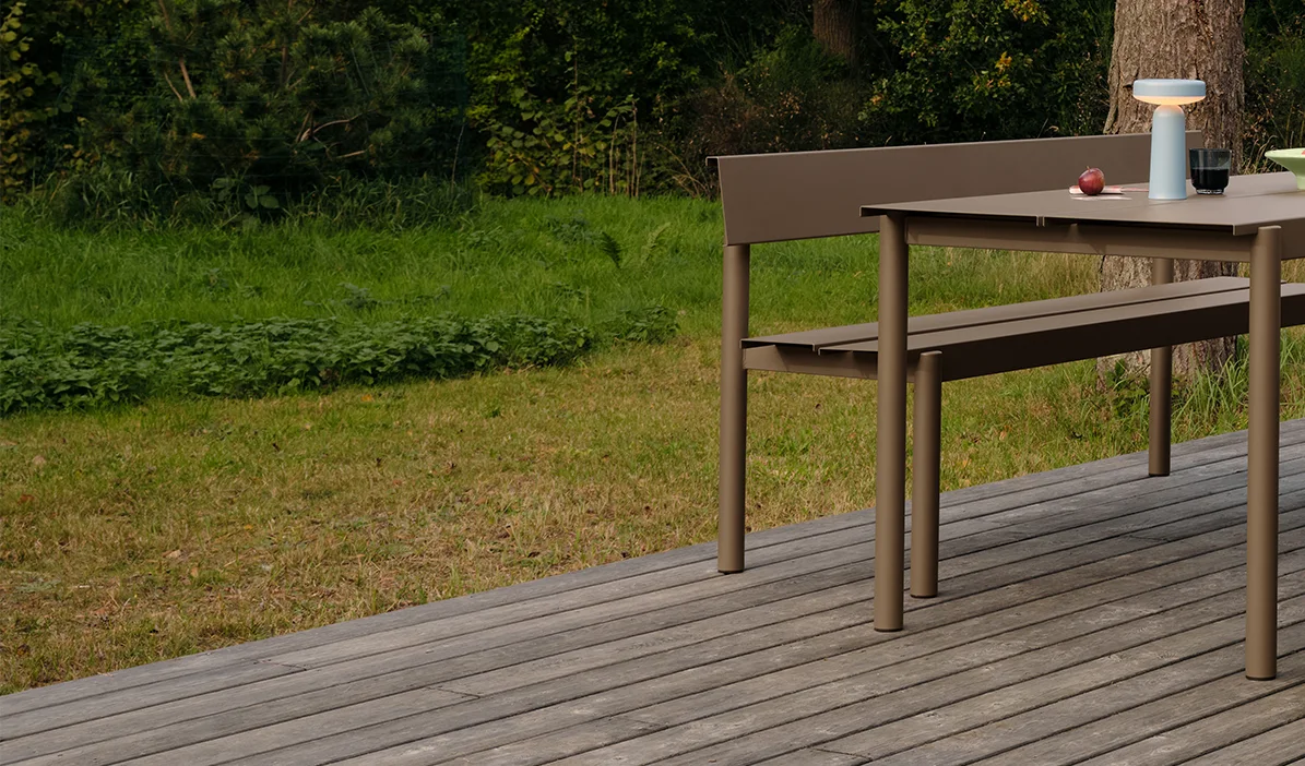 Modern outdoor dining table and bench in taupe on a wooden deck, with a portable lamp and apples on the table.