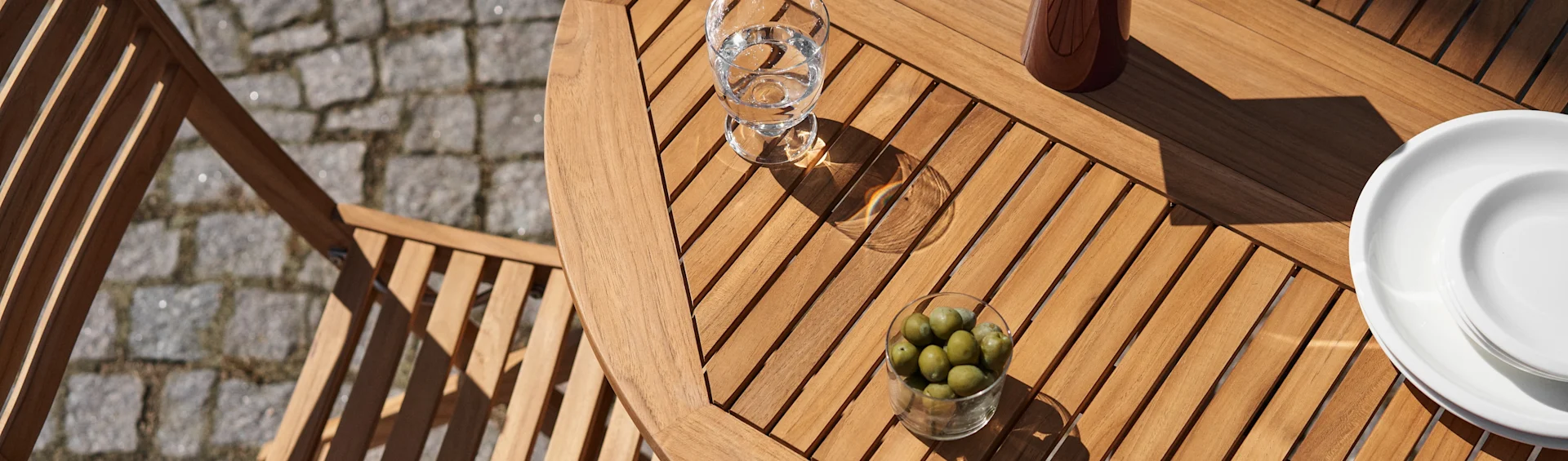 Overhead view of a wooden slatted patio table with a glass of water, a bowl of green olives, and white plates in sunlight.