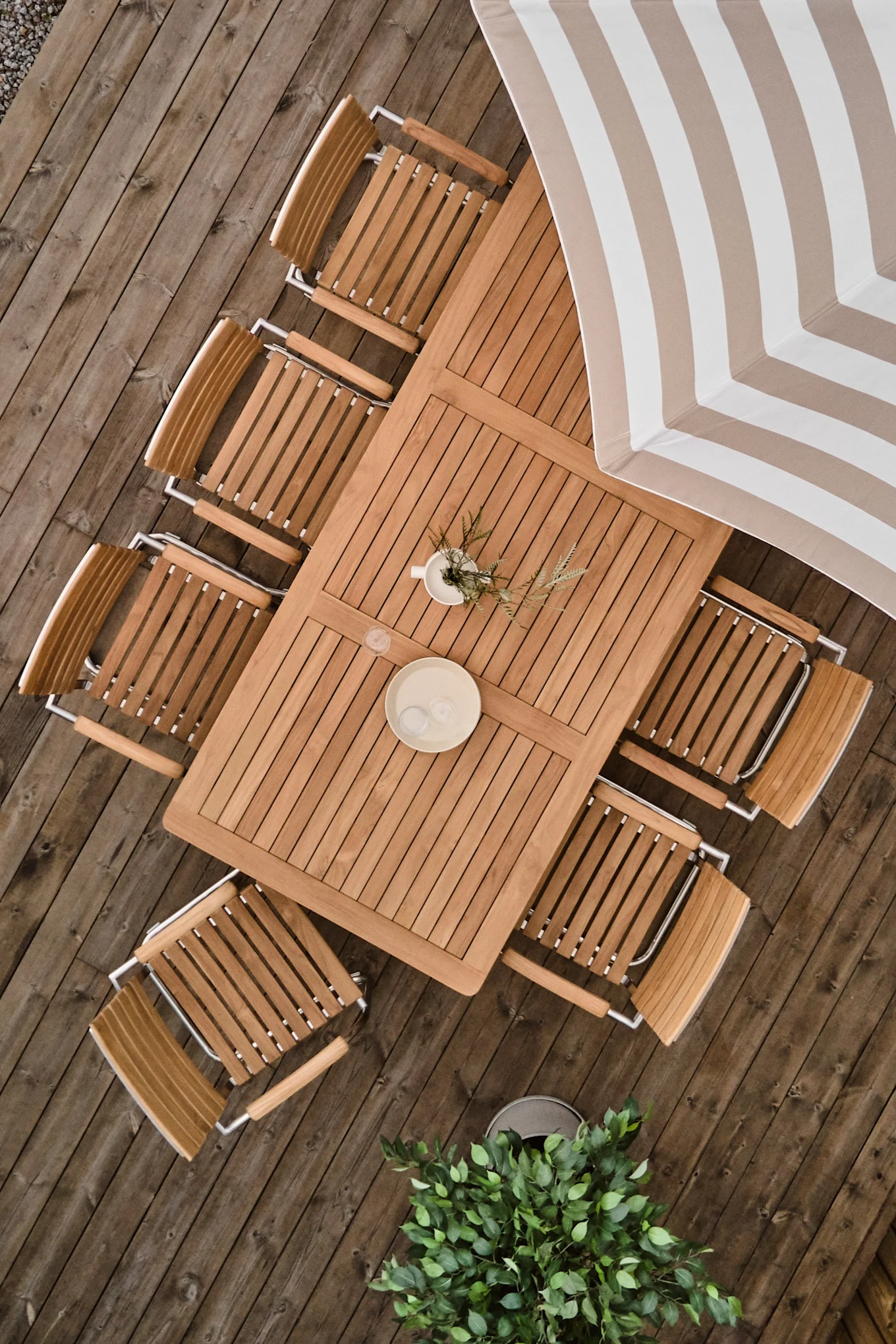 Overhead view of a rectangular wooden patio dining table with eight chairs on a wooden deck, a striped umbrella, and potted plants.