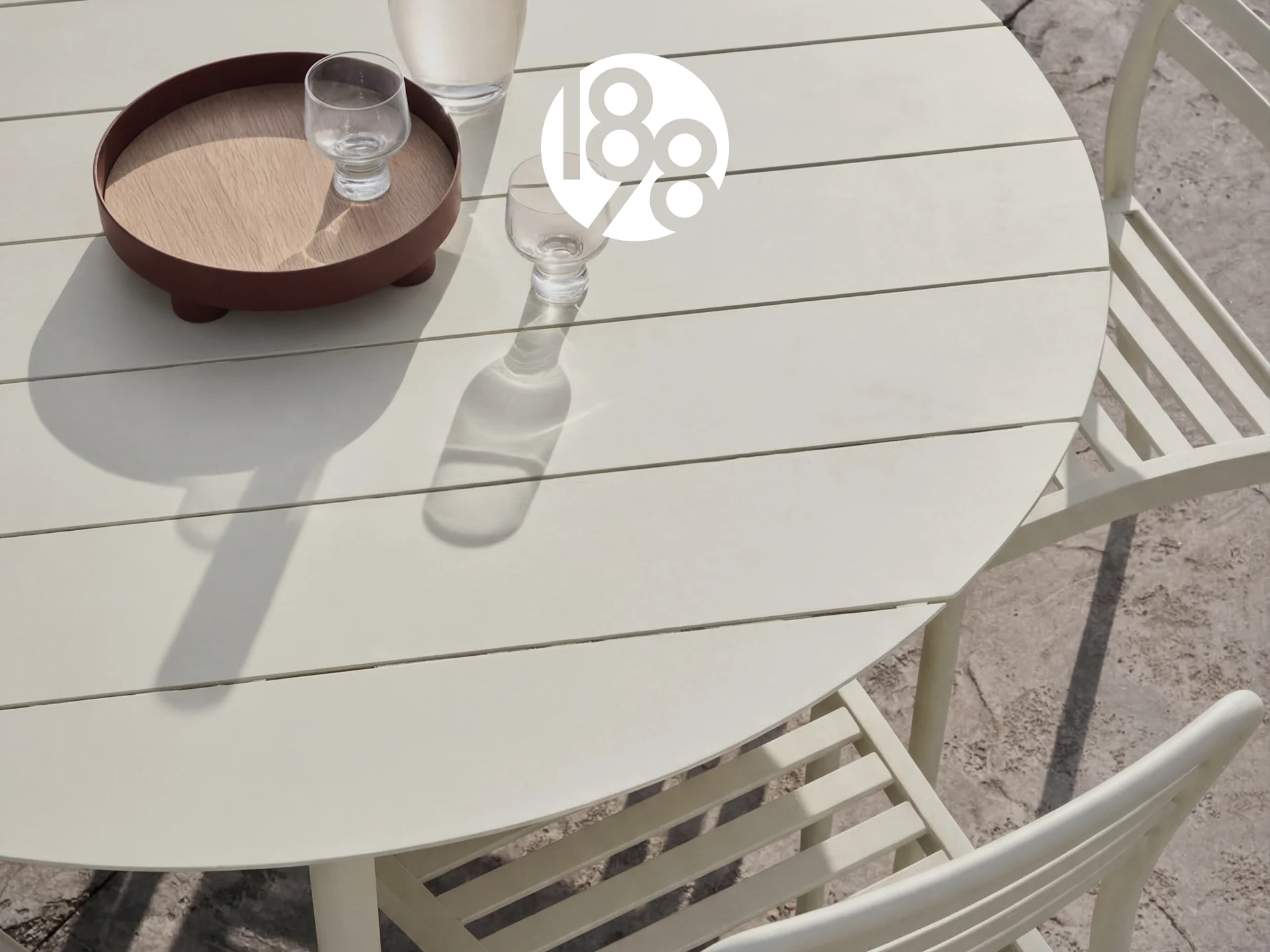 Outdoor round light beige slatted table with two chairs, a tray, and glasses on a patio.