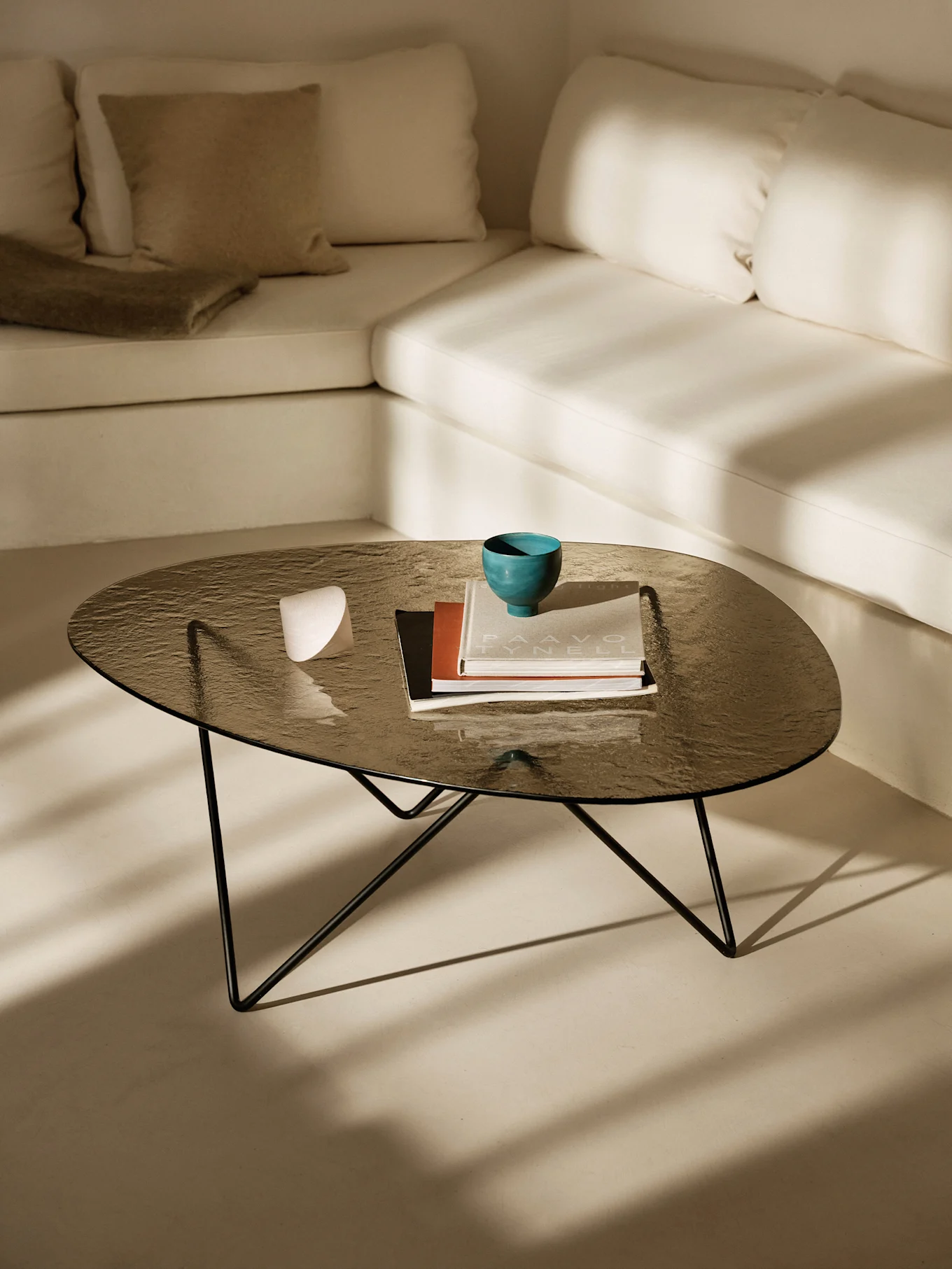 Dark glass coffee table with a teal bowl, books, and a white object. Light L-shaped sofa and sunlight shadows.