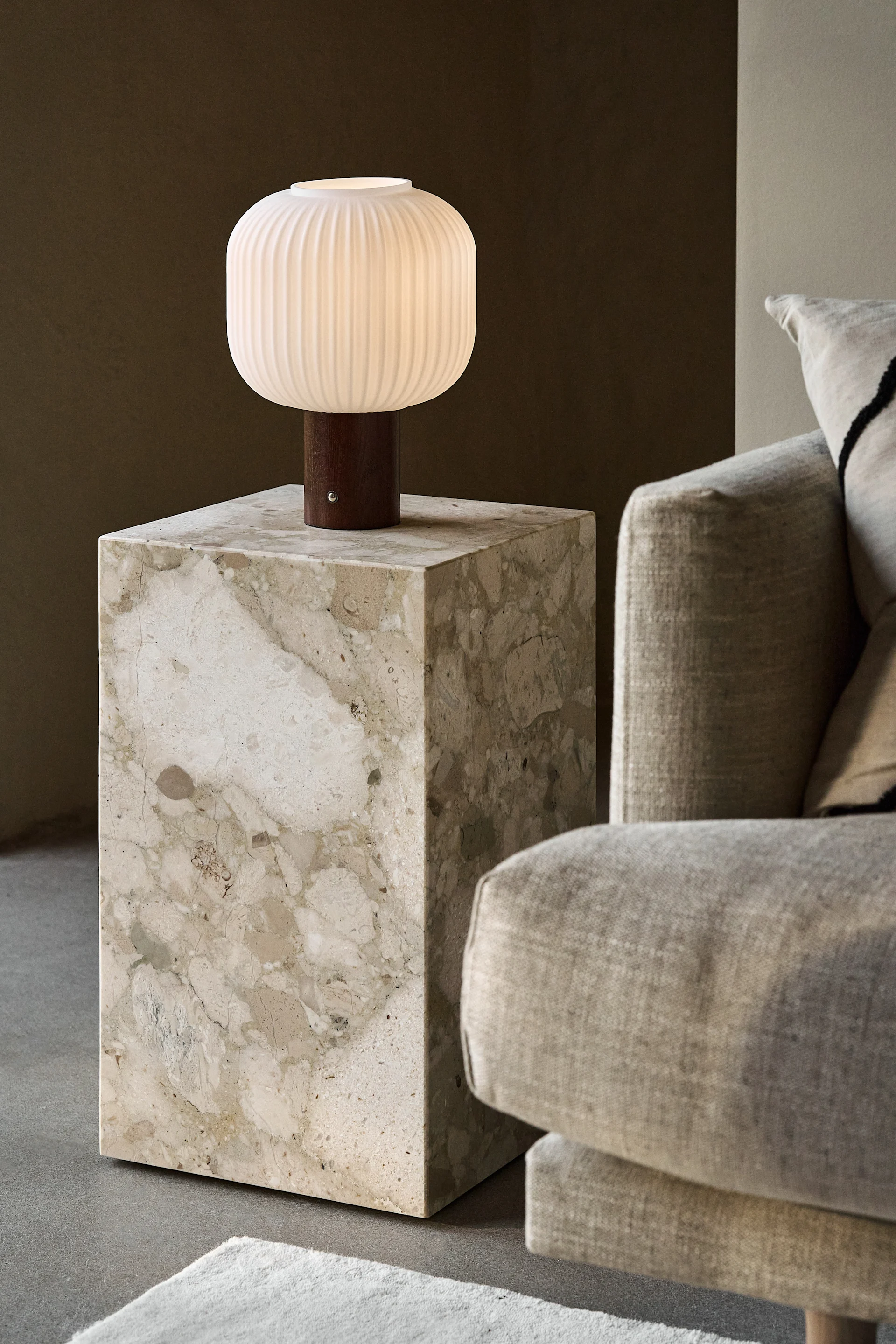 A lit, ribbed, white table lamp with a wooden base on a beige marble side table next to a light-colored sofa.