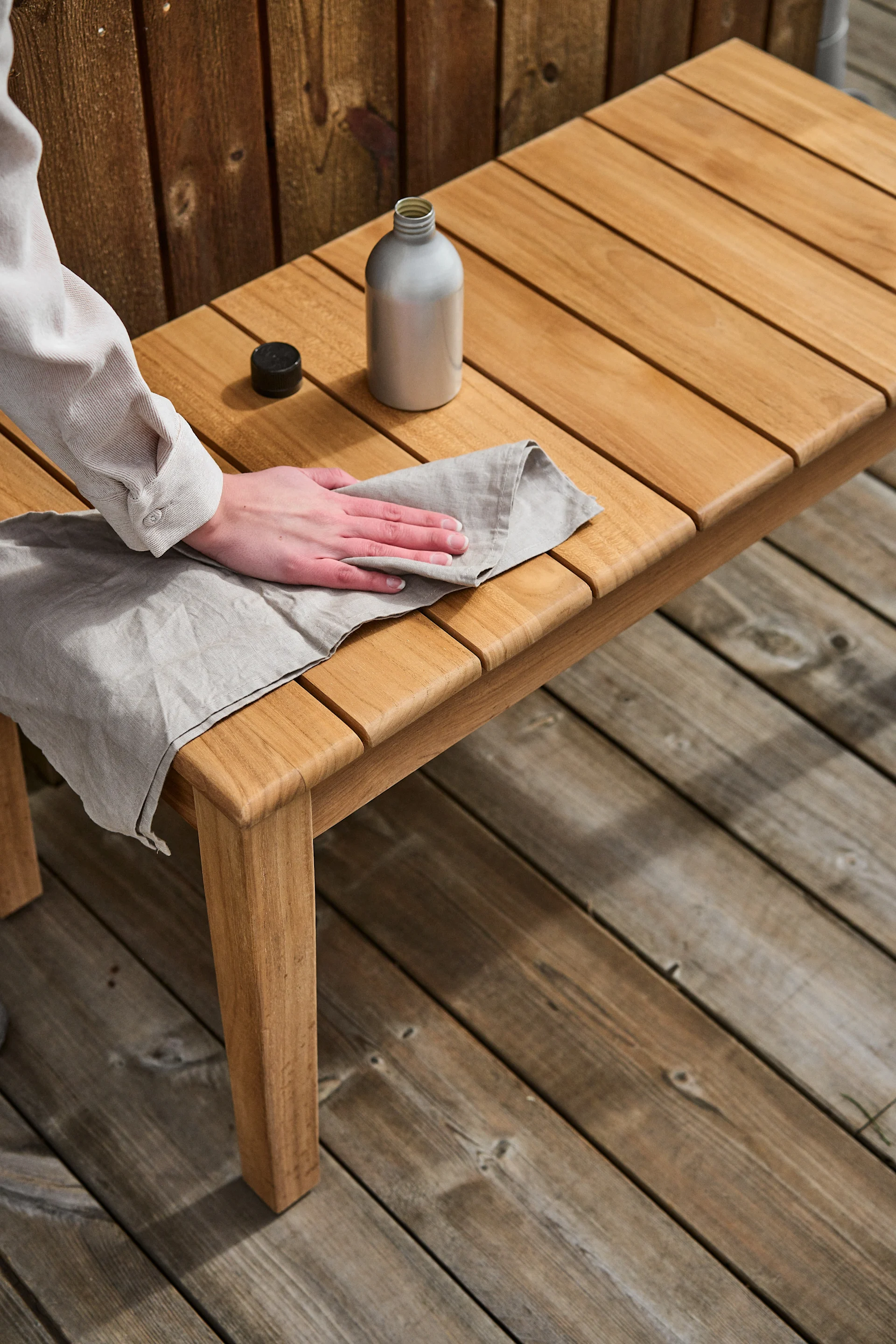 A pair of hands apply oil to a cloth in order to apply teak oil to a teak wood bench.