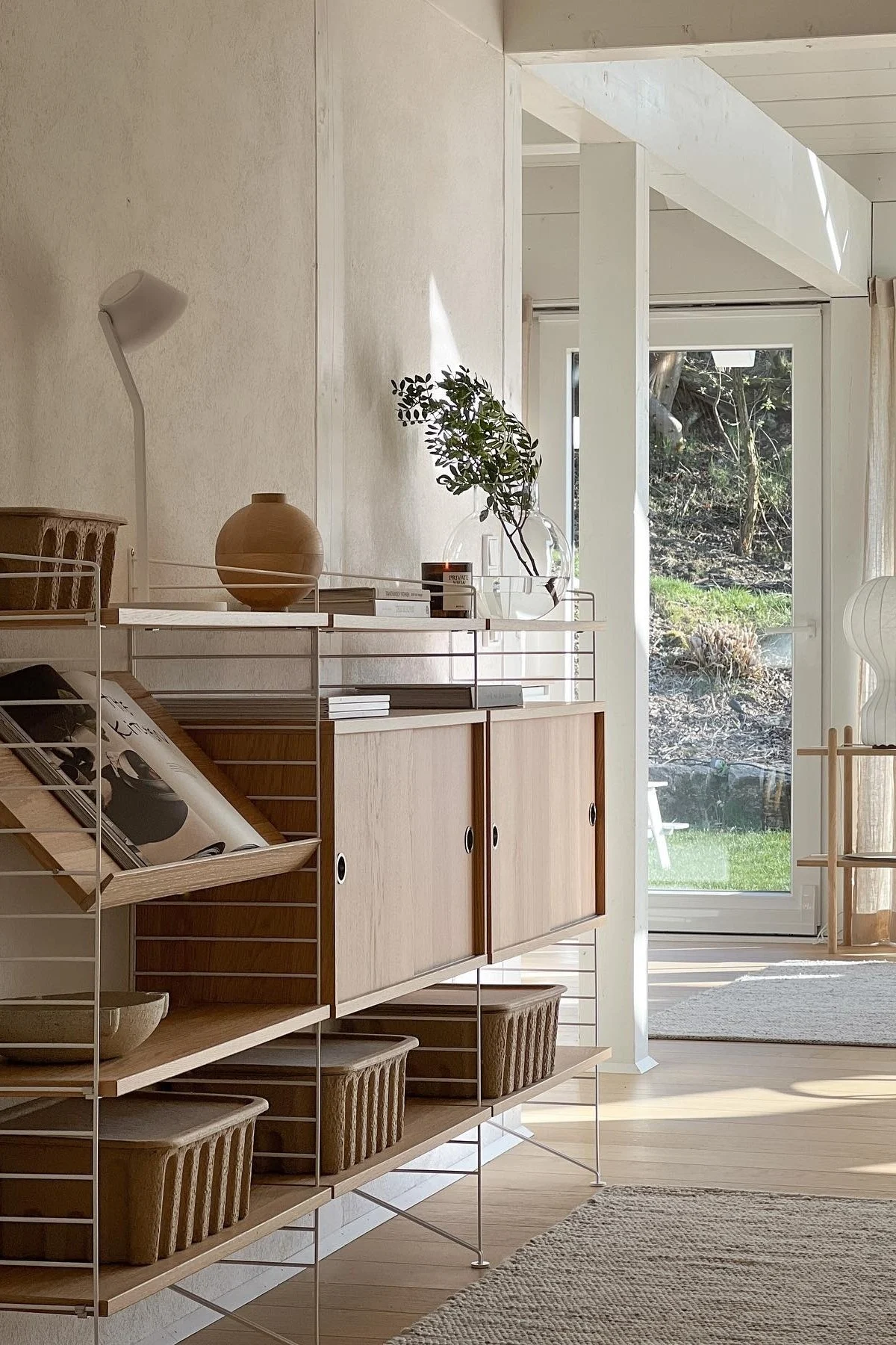 A sunny room with a modern modular shelving unit made of light wood and white frames, holding a wooden vase, books, greenery, and woven baskets, with a garden view outside.