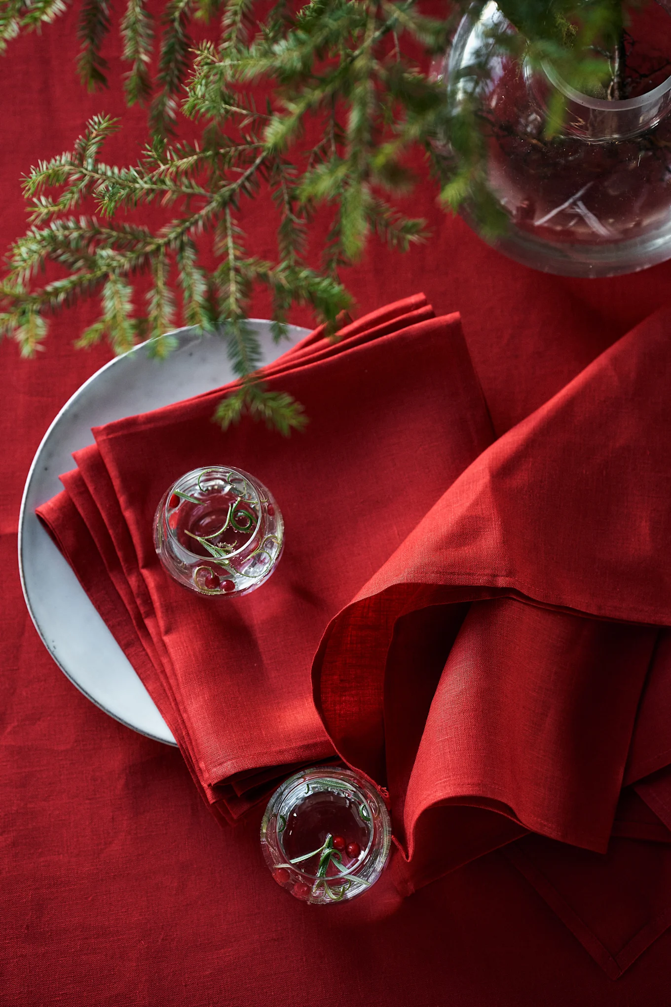 Festive red table setting with stacked red linen napkins on a plate, small glass bowls with cranberries and pine branches.