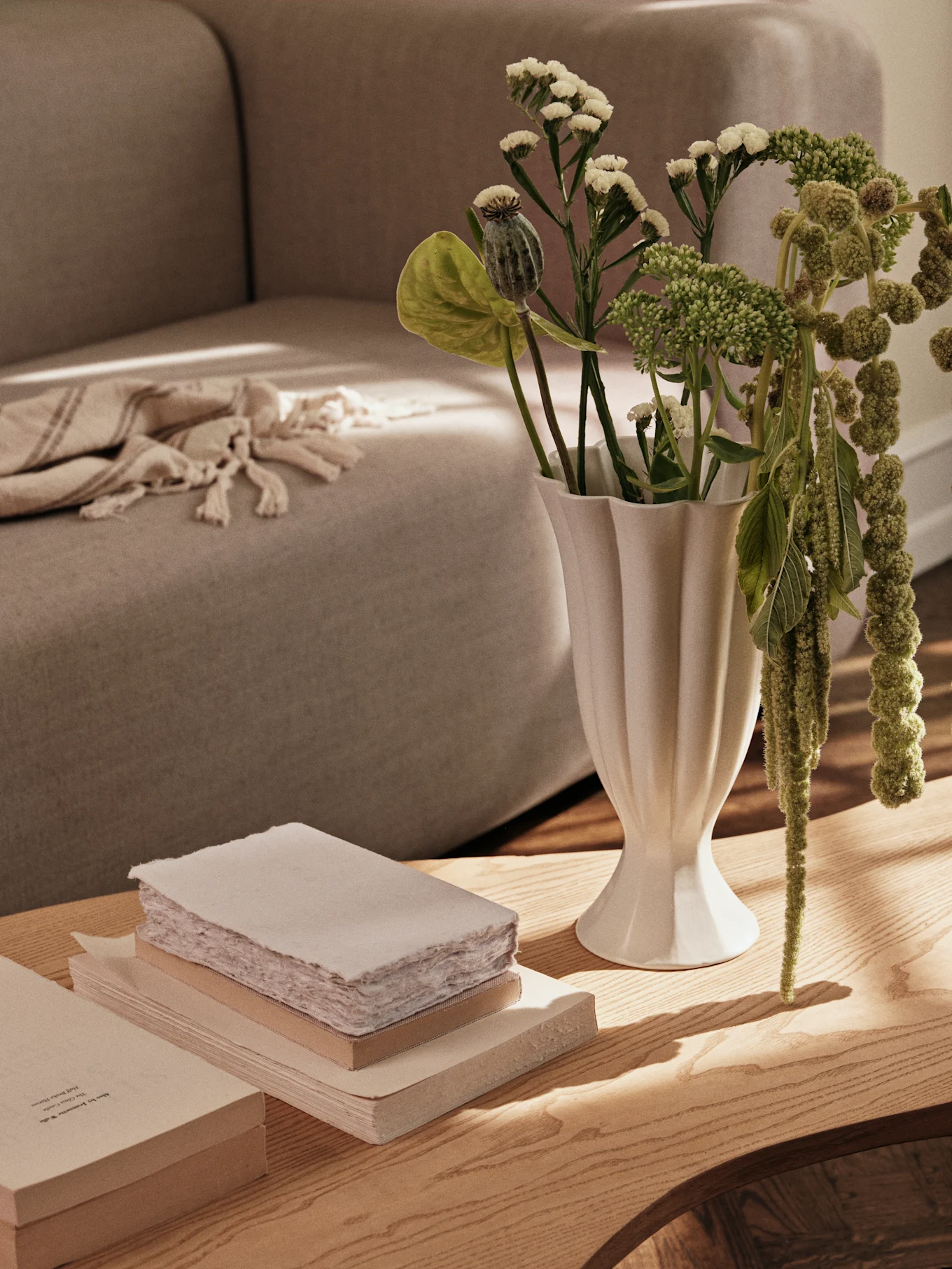 A white fluted vase with green plants and a stack of books on a wooden coffee table, with a sofa in the background.