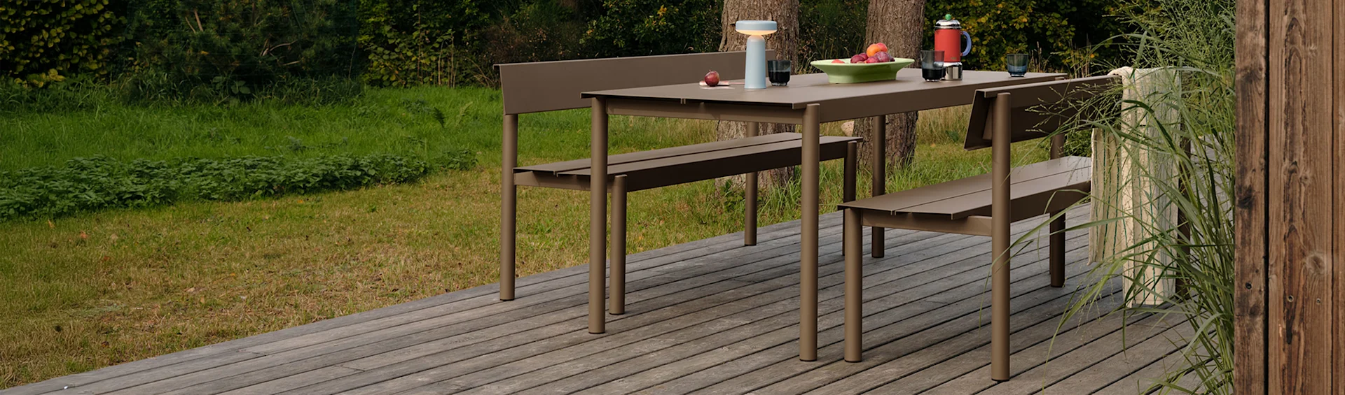 Modern taupe outdoor dining set: table, two benches, fruit, lamp, coffee pot on wooden deck, surrounded by greenery.