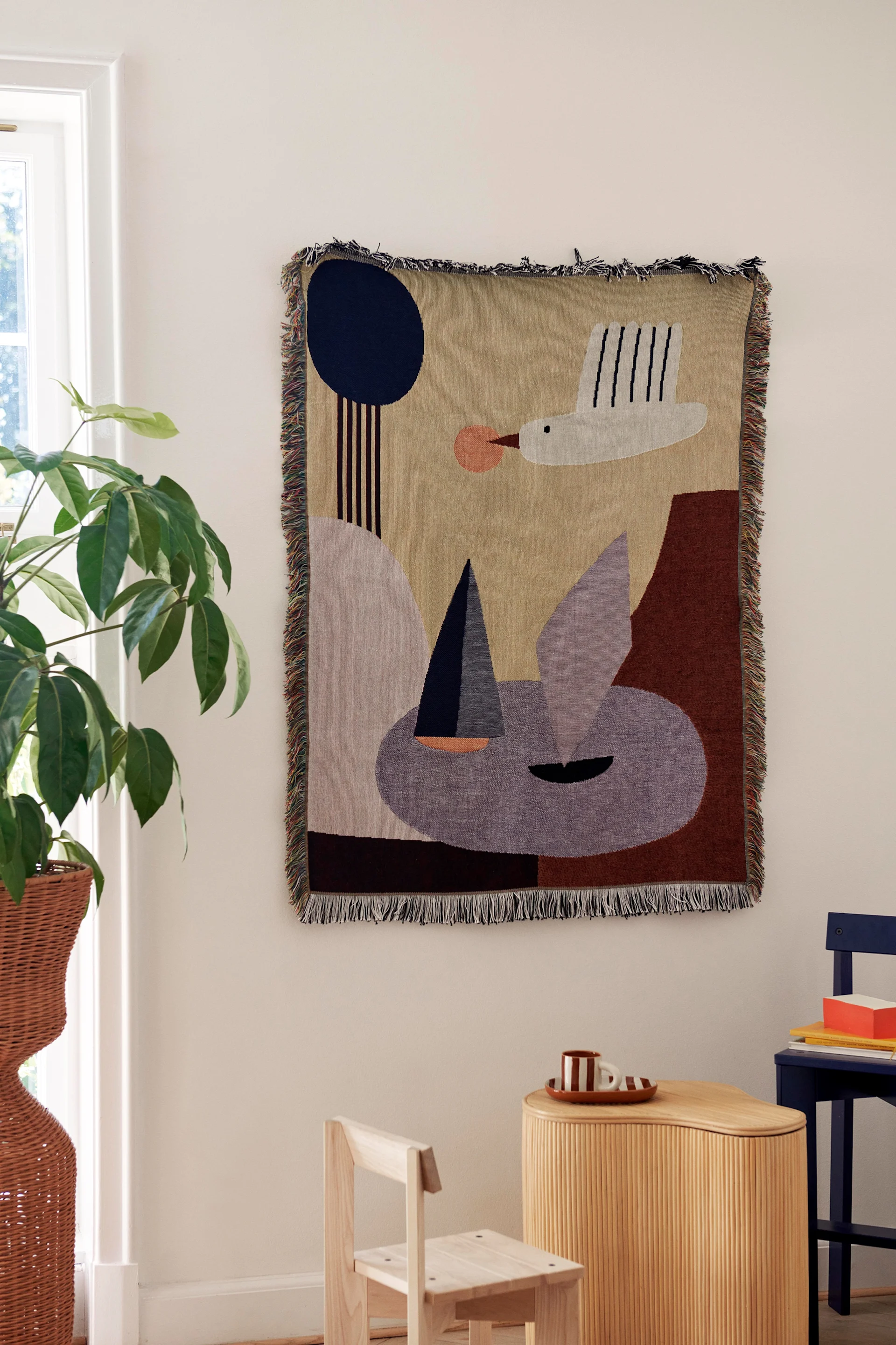 Abstract tapestry with bird, sailboat, and geometric shapes in earthy tones on white wall, with a plant and wooden furniture.