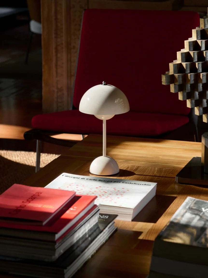 A white mushroom table lamp, books, and a geometric sculpture on a wooden coffee table, with a red armchair behind.