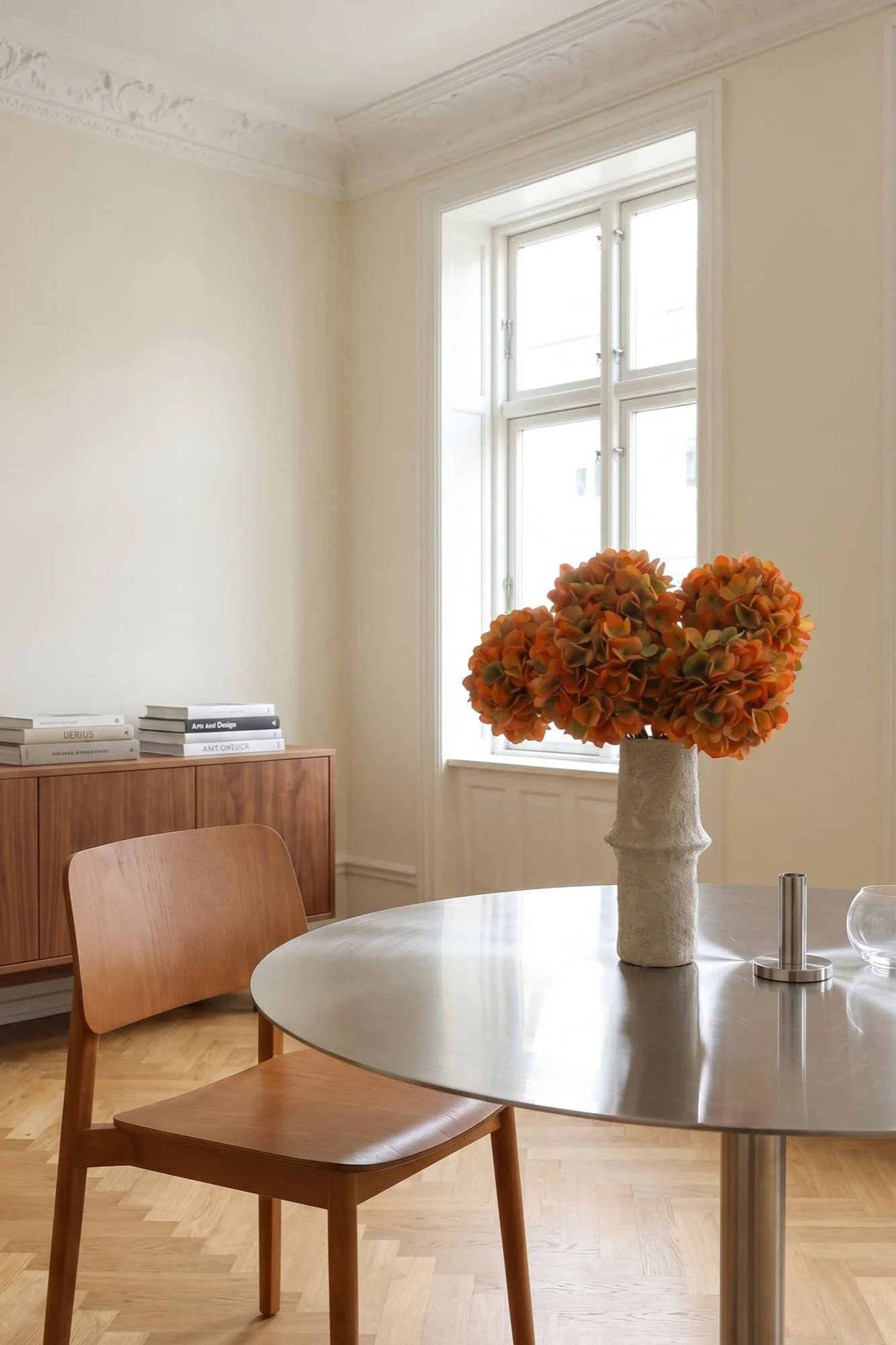 A bright dining room with a round metal table, a wooden chair, and a vase of orange flowers.