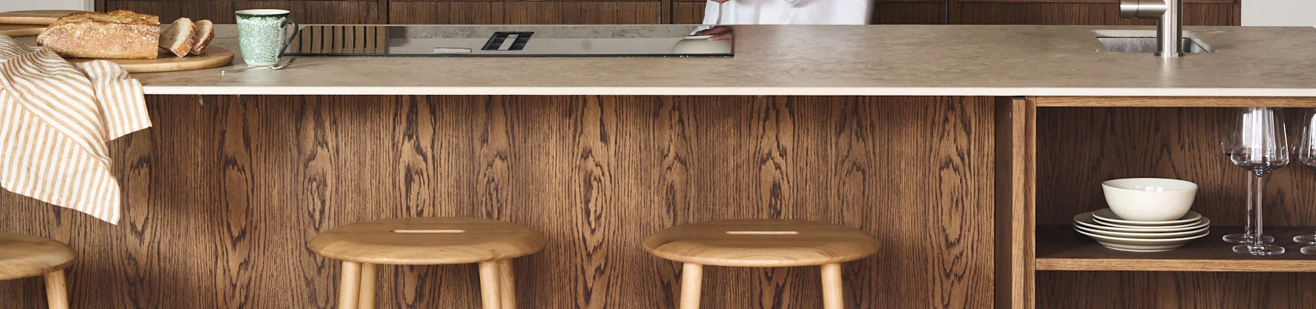 Modern kitchen island with light countertop, integrated hob, sink, fresh bread, a mug, and two wooden stools against a dark wood base.