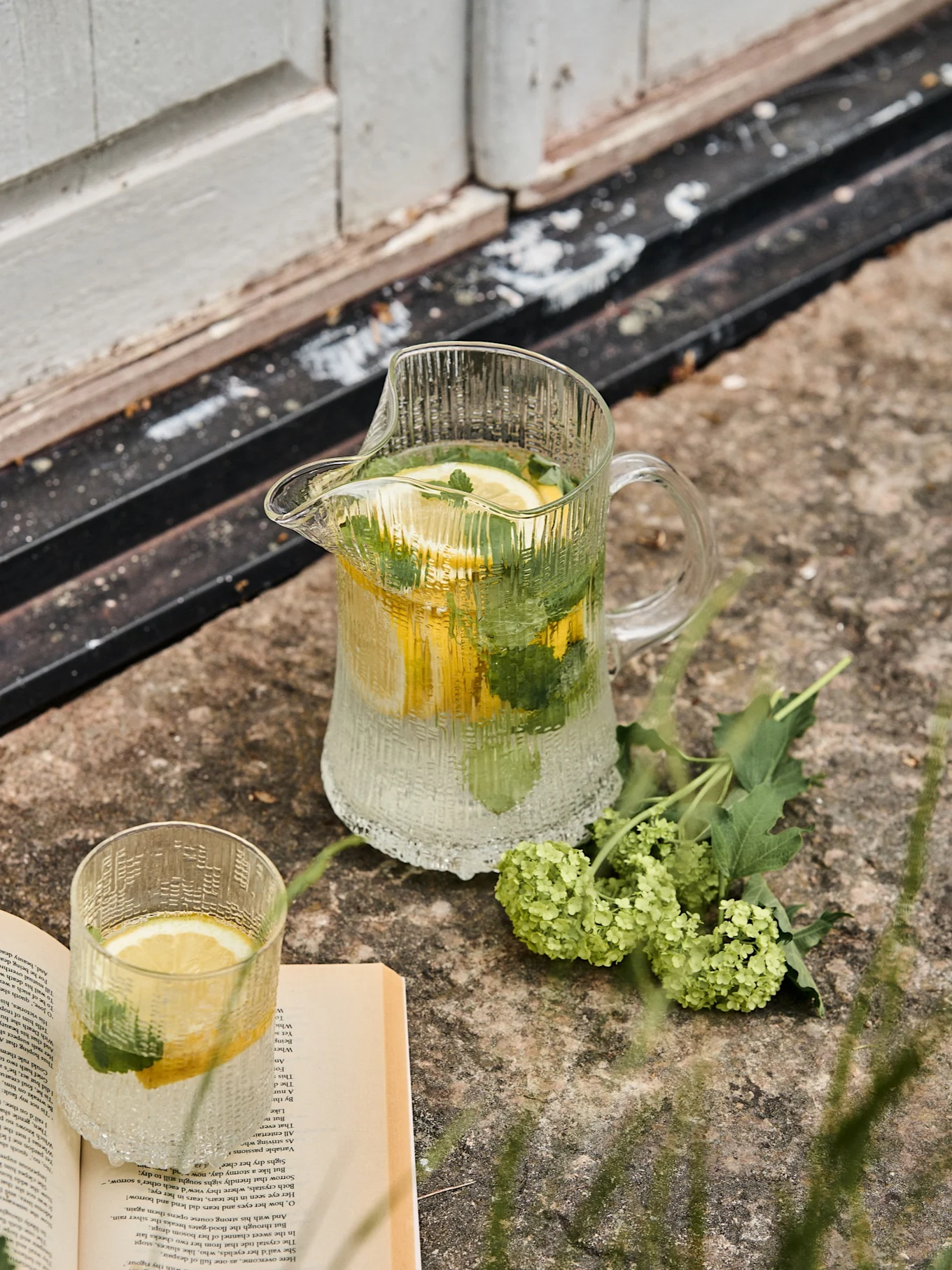 Ribbed glass pitcher and matching glass with lemon and mint water, next to an open book and green flowers on stone.