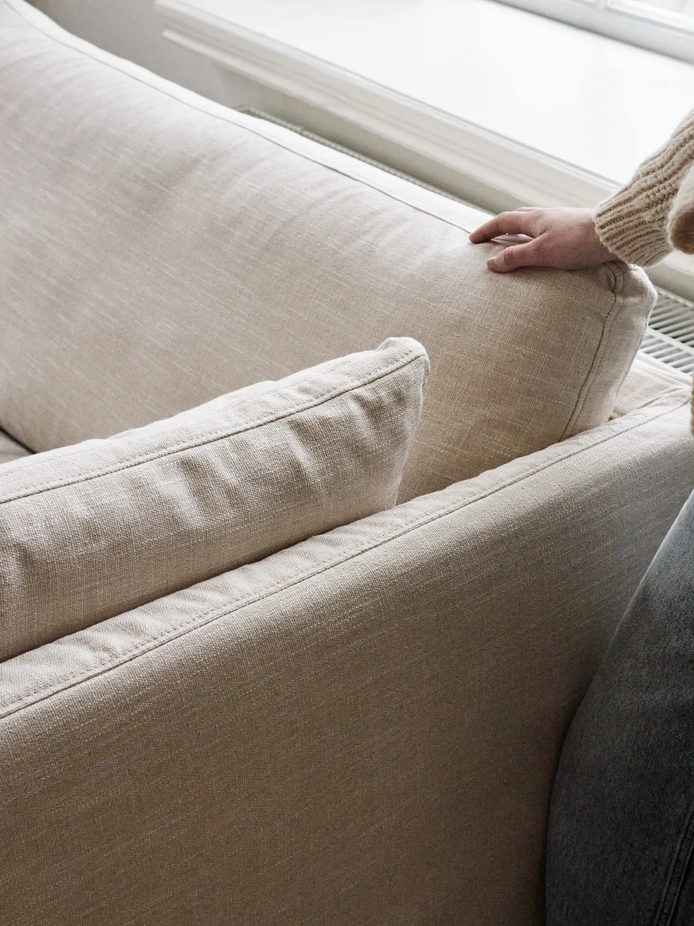 A hand in a chunky knit sweater gently presses into the back cushion of a light beige sofa.