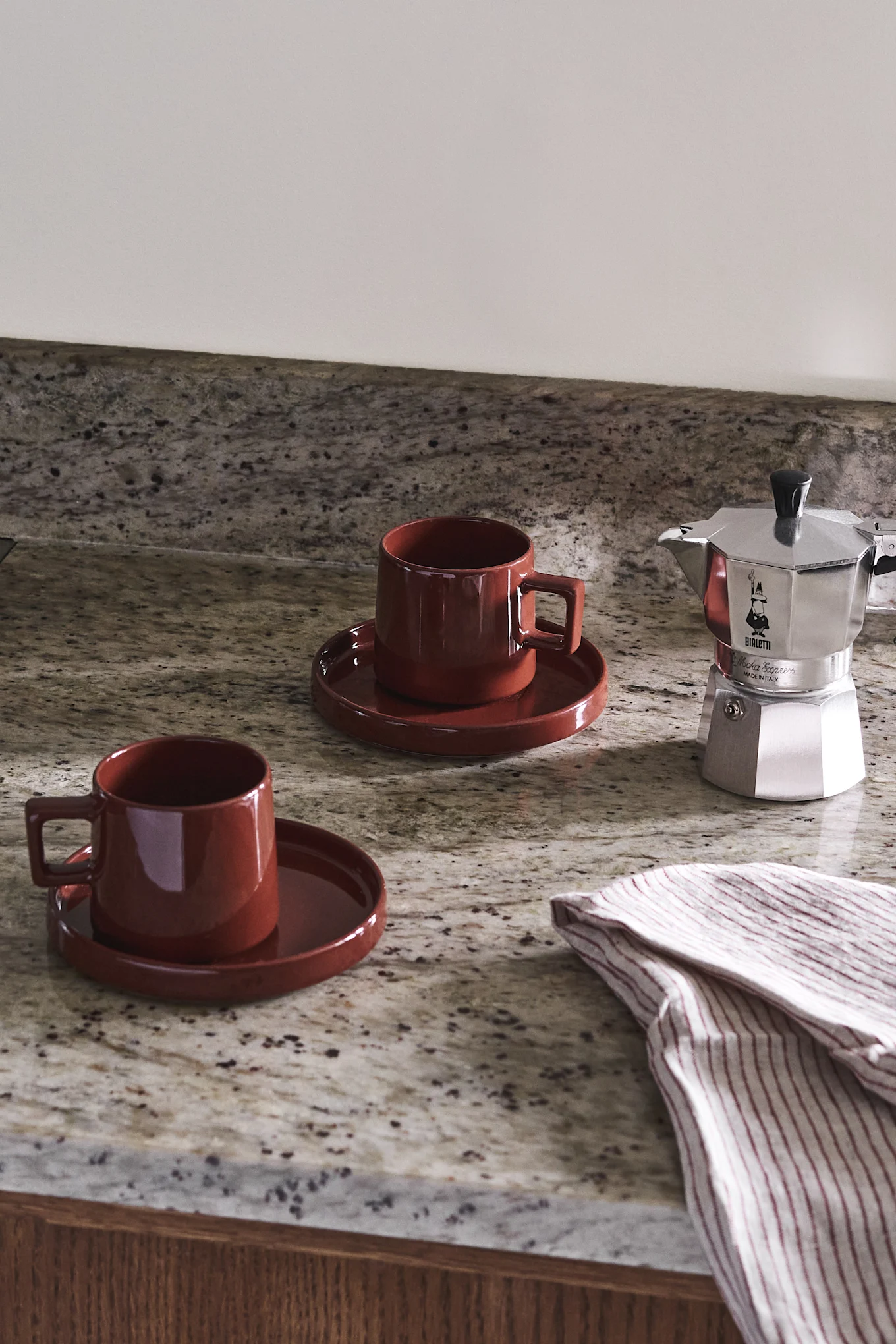 Two red ceramic mugs and a silver coffee maker on a speckled granite countertop with a striped towel.