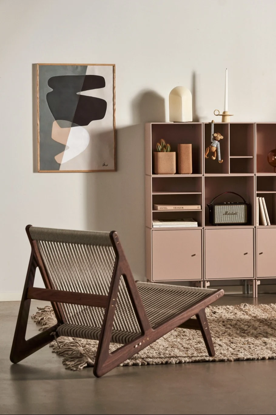 A modern interior features a wooden lounge chair with rope weaving, a pink modular cabinet, and abstract wall art.