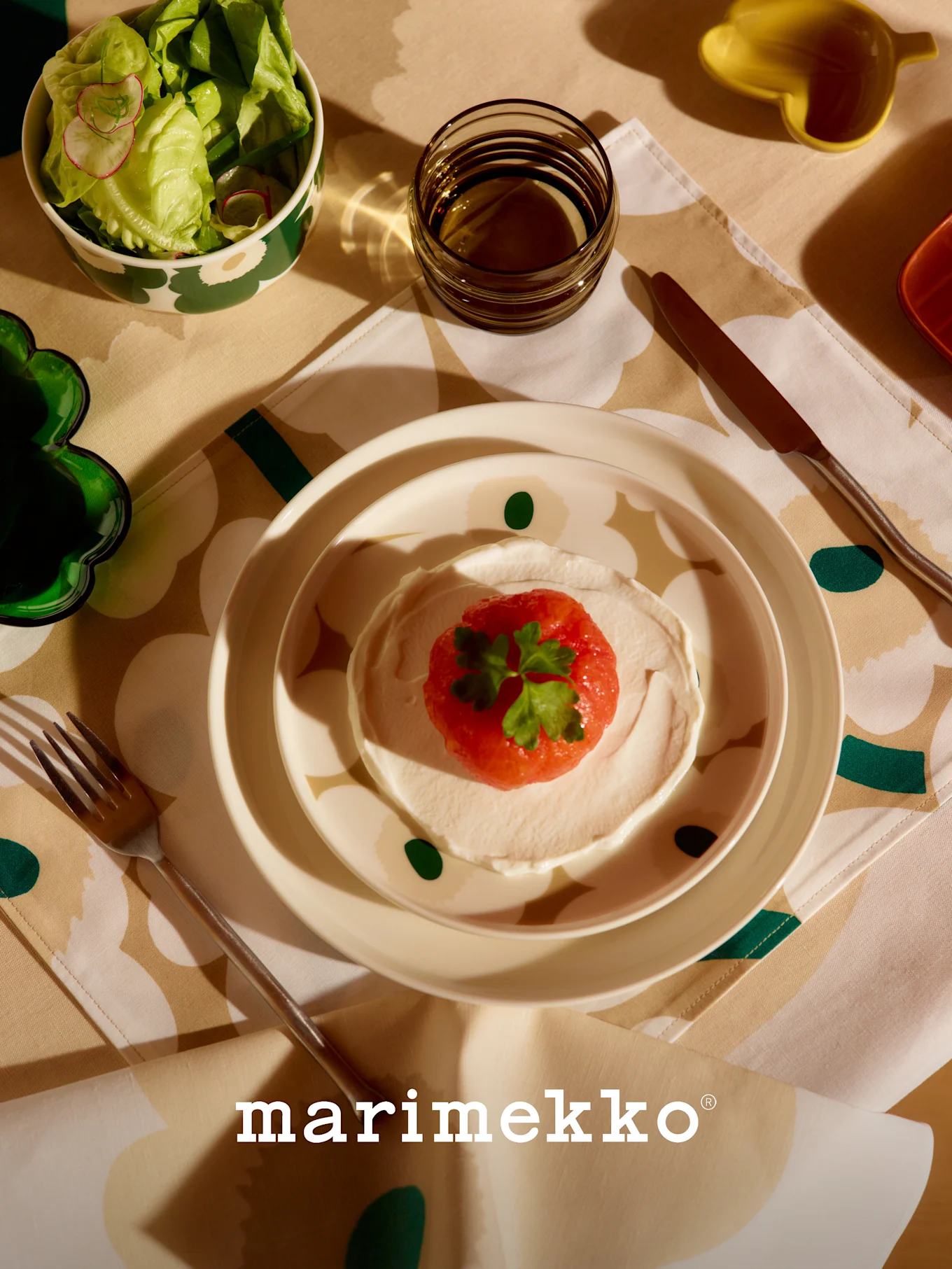 Marimekko table setting with patterned salad bowl, red fruit and cream dish, glass, and cutlery on a floral tablecloth.
