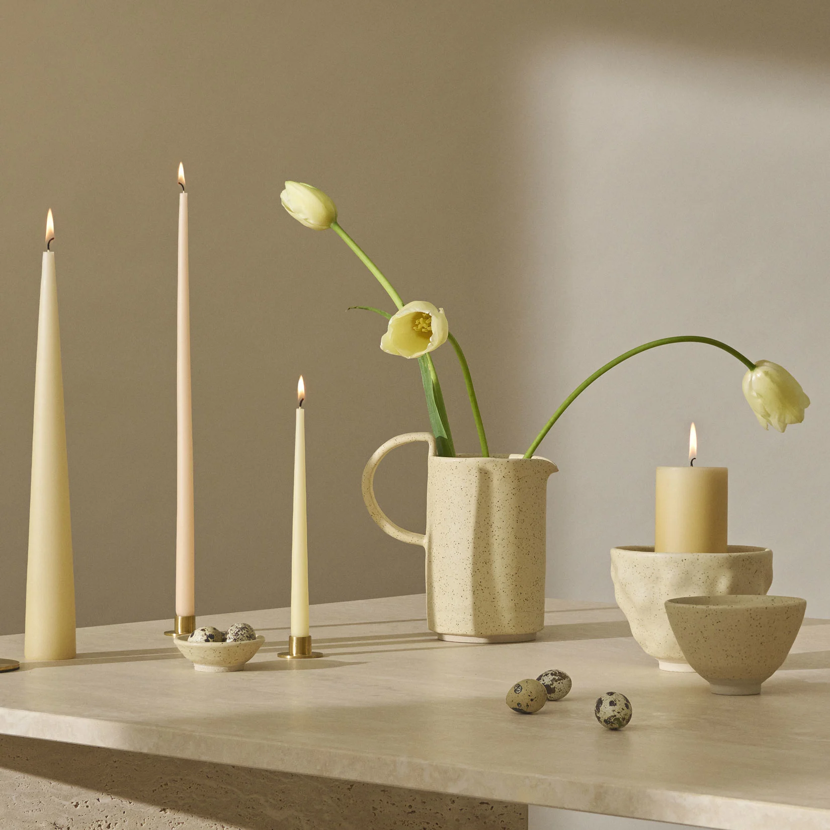 A still life with lit candles, pale yellow tulips in a speckled pitcher, and quail eggs on a textured surface.
