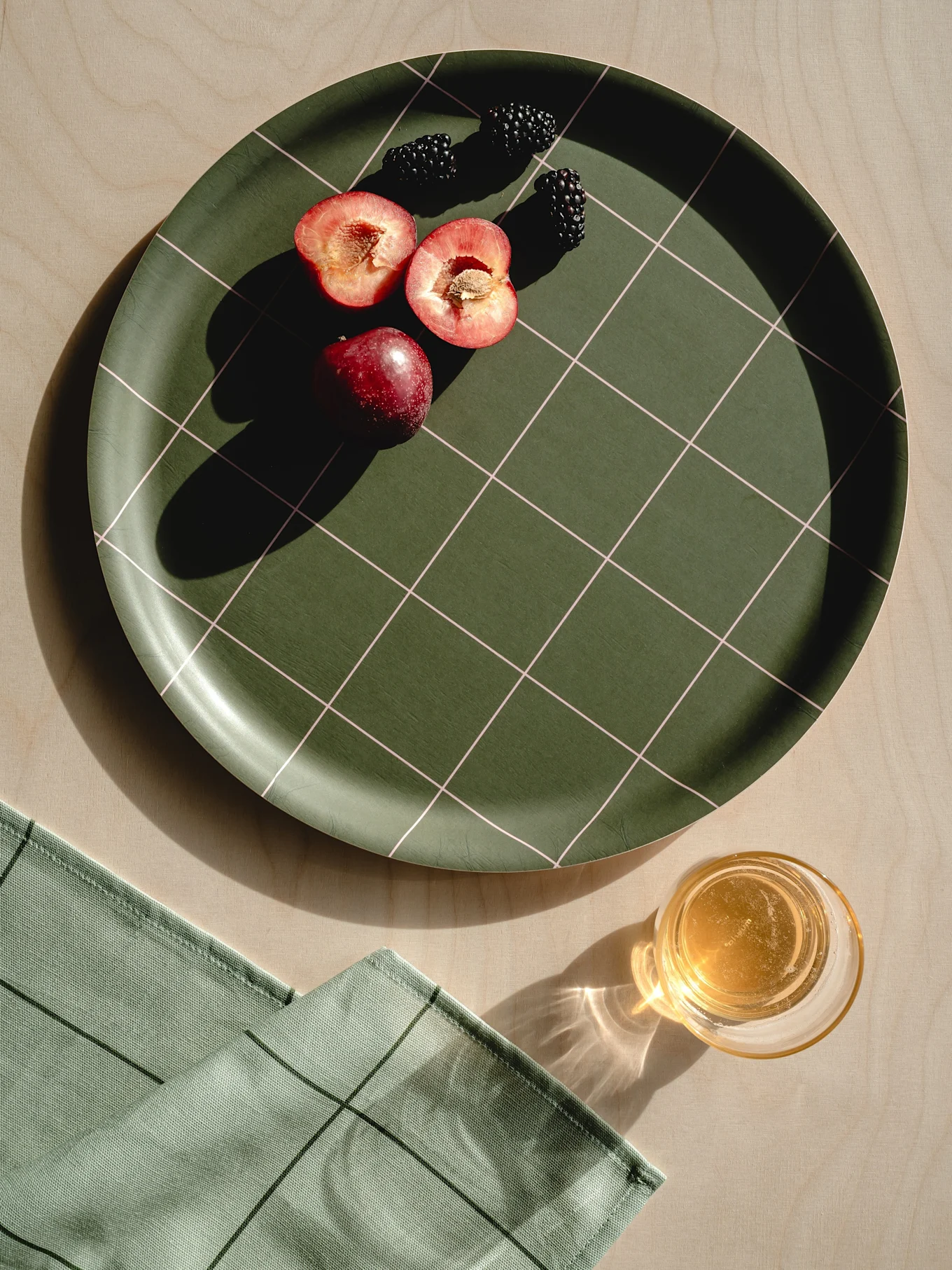 Dark green checkered tray with red plums, blackberries, light green napkin, and a glass of juice on wood.