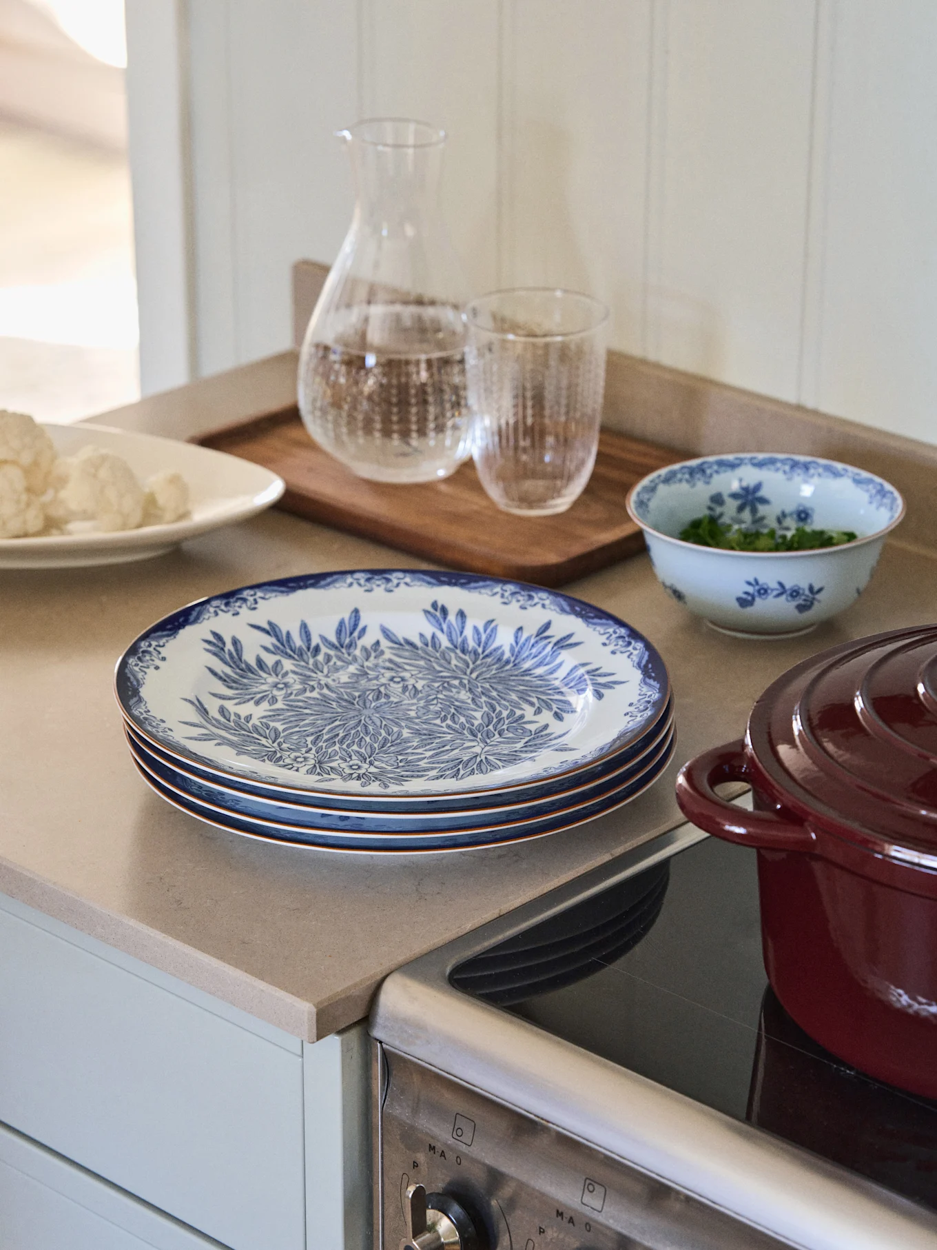 A kitchen counter features a stack of blue and white patterned plates, a bowl of cauliflower, a glass carafe of water, and a red pot on a cooktop.