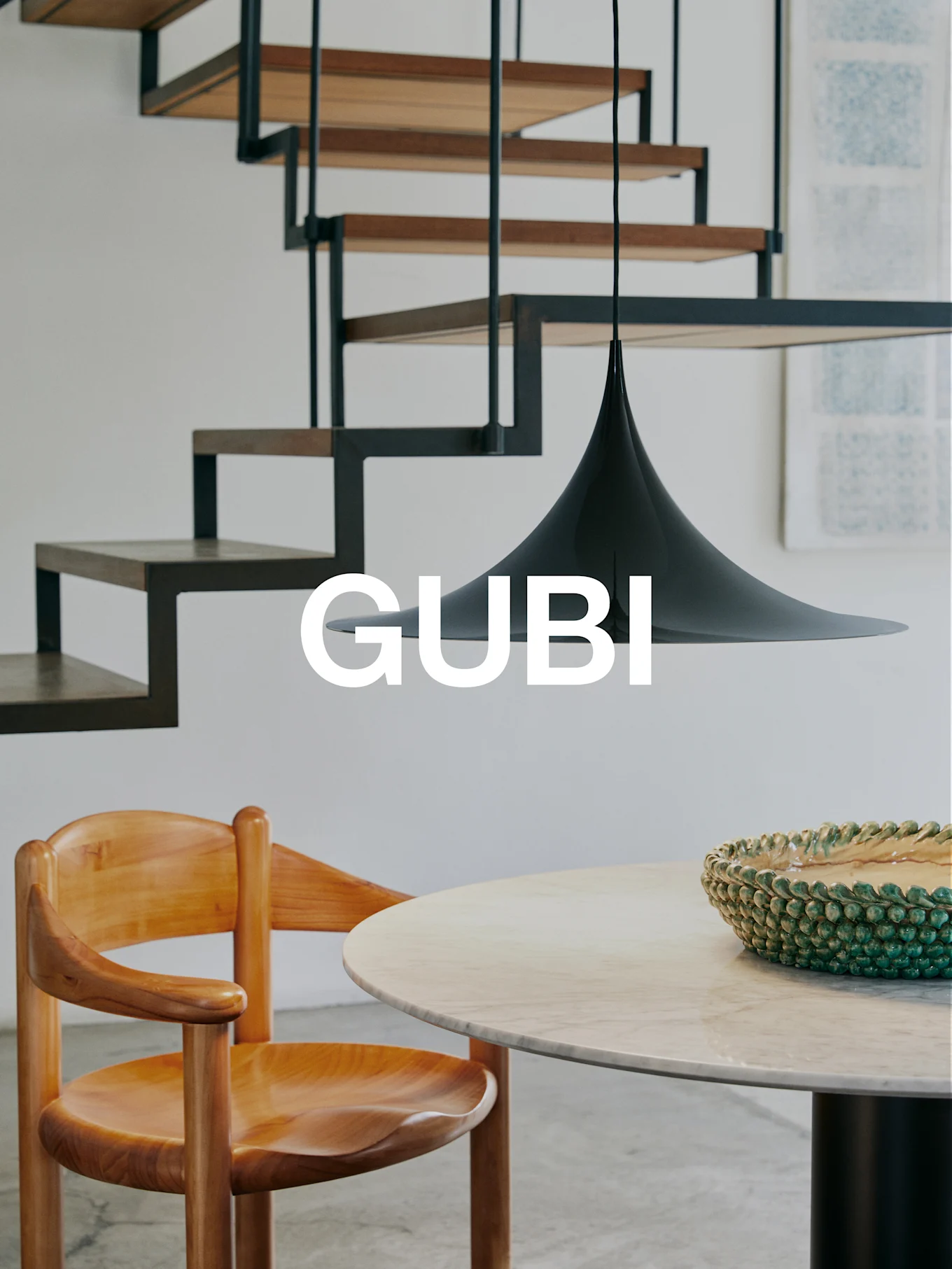 GUBI logo in a modern interior with a wooden chair, marble table, green bowl, black pendant lamp, and a modern staircase.