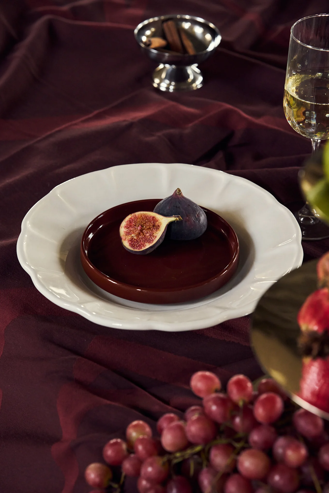 Figs on a plate on a burgundy tablecloth with wine, grapes, and cinnamon sticks.