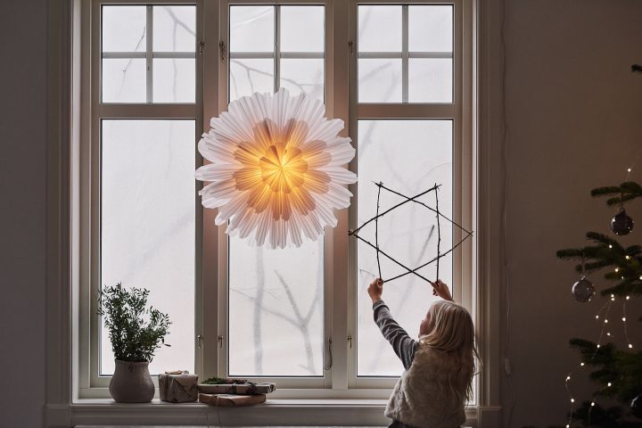Snöblomma Christmas star from Watt & Veke in a window with a child holding a star made from of sticks.