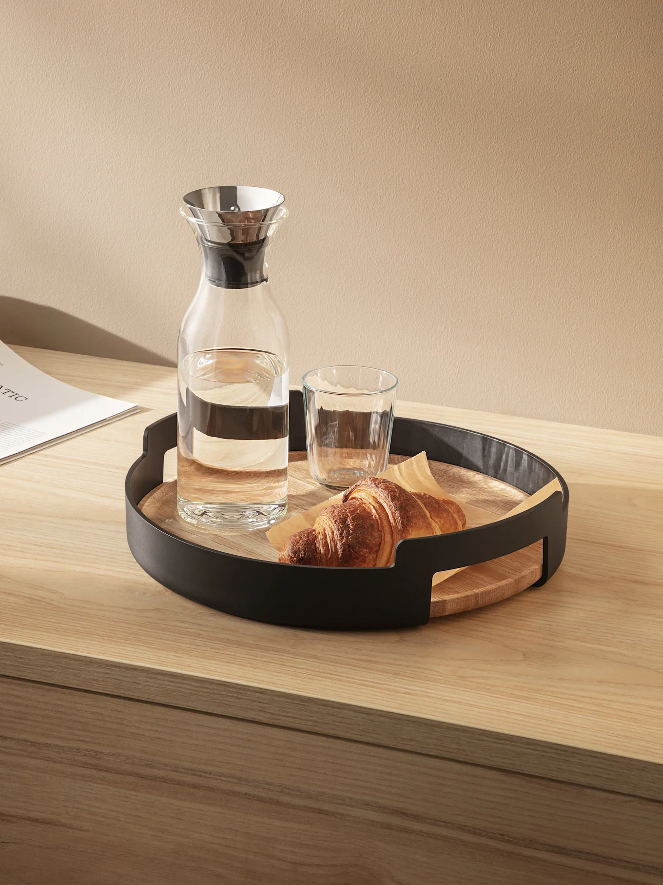 A glass carafe of water, a glass, and a croissant on a round wooden tray with a black metal rim, on a light wooden table.