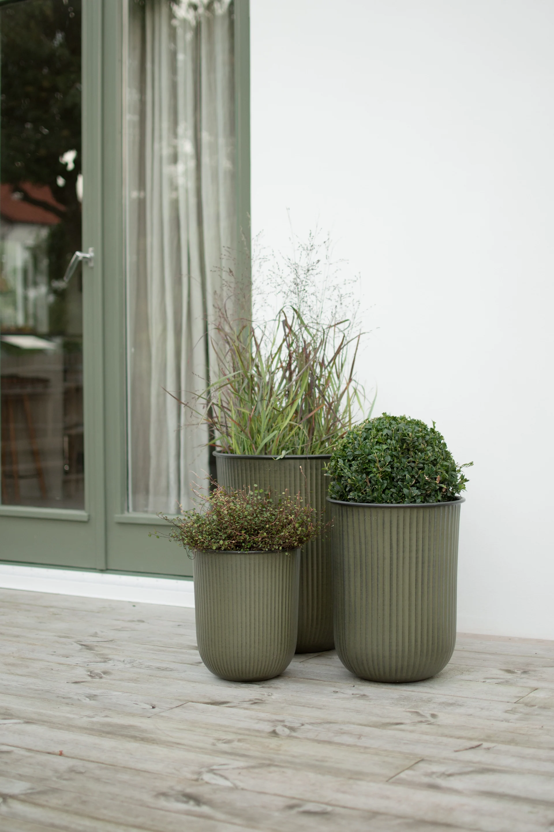 Three olive green ribbed planters of varying heights with different plants, on a light wooden deck next to a green door frame.