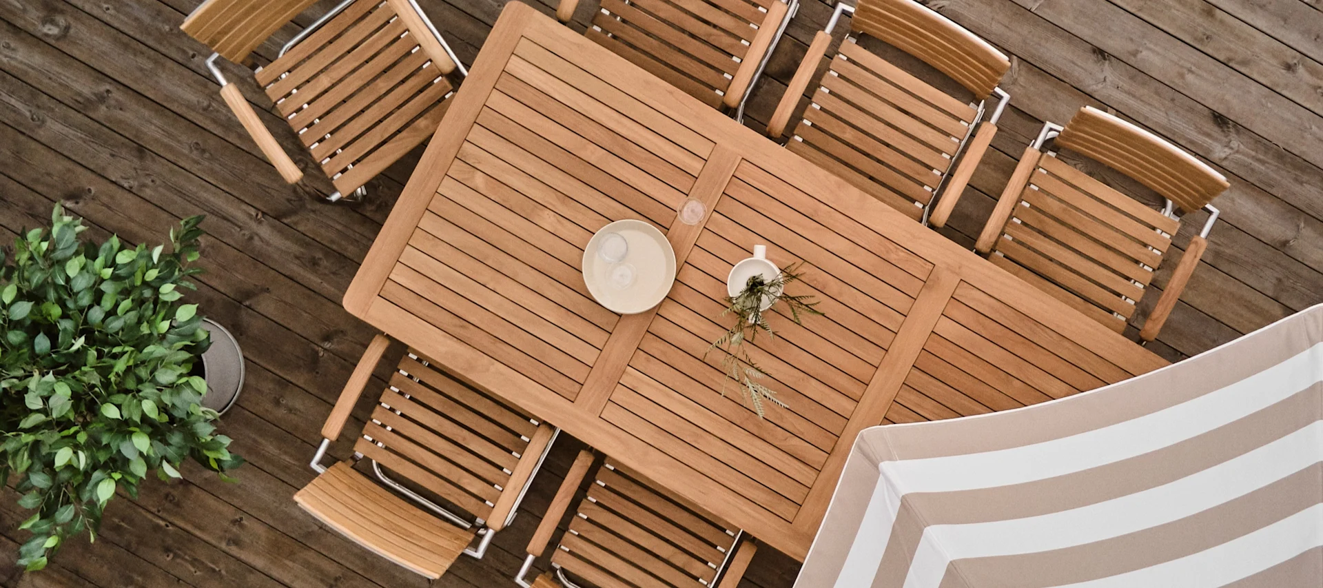 Overhead view: a wooden outdoor dining table with chairs on a deck. A potted plant is on the left, and a striped umbrella is on the right.