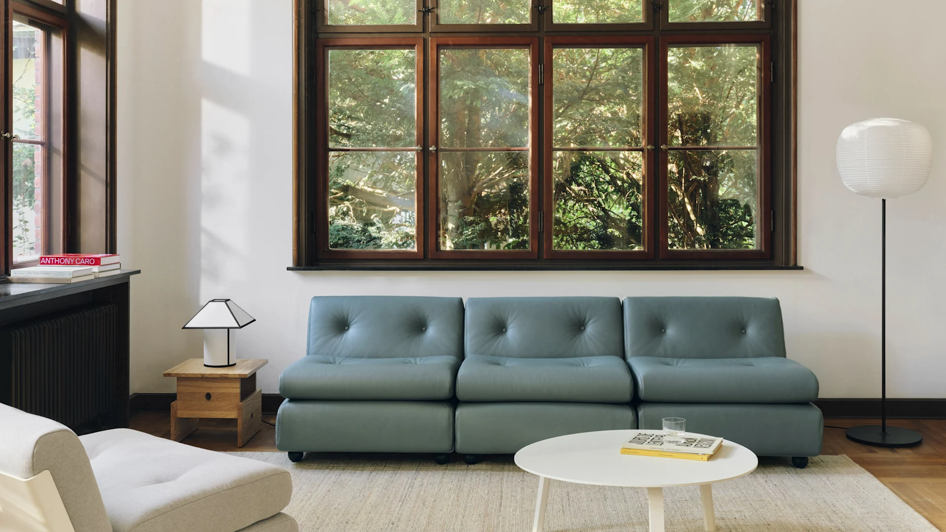 A modern living room with a blue-grey modular sofa, white coffee table, and a large window overlooking green trees. Two lamps illuminate the space.