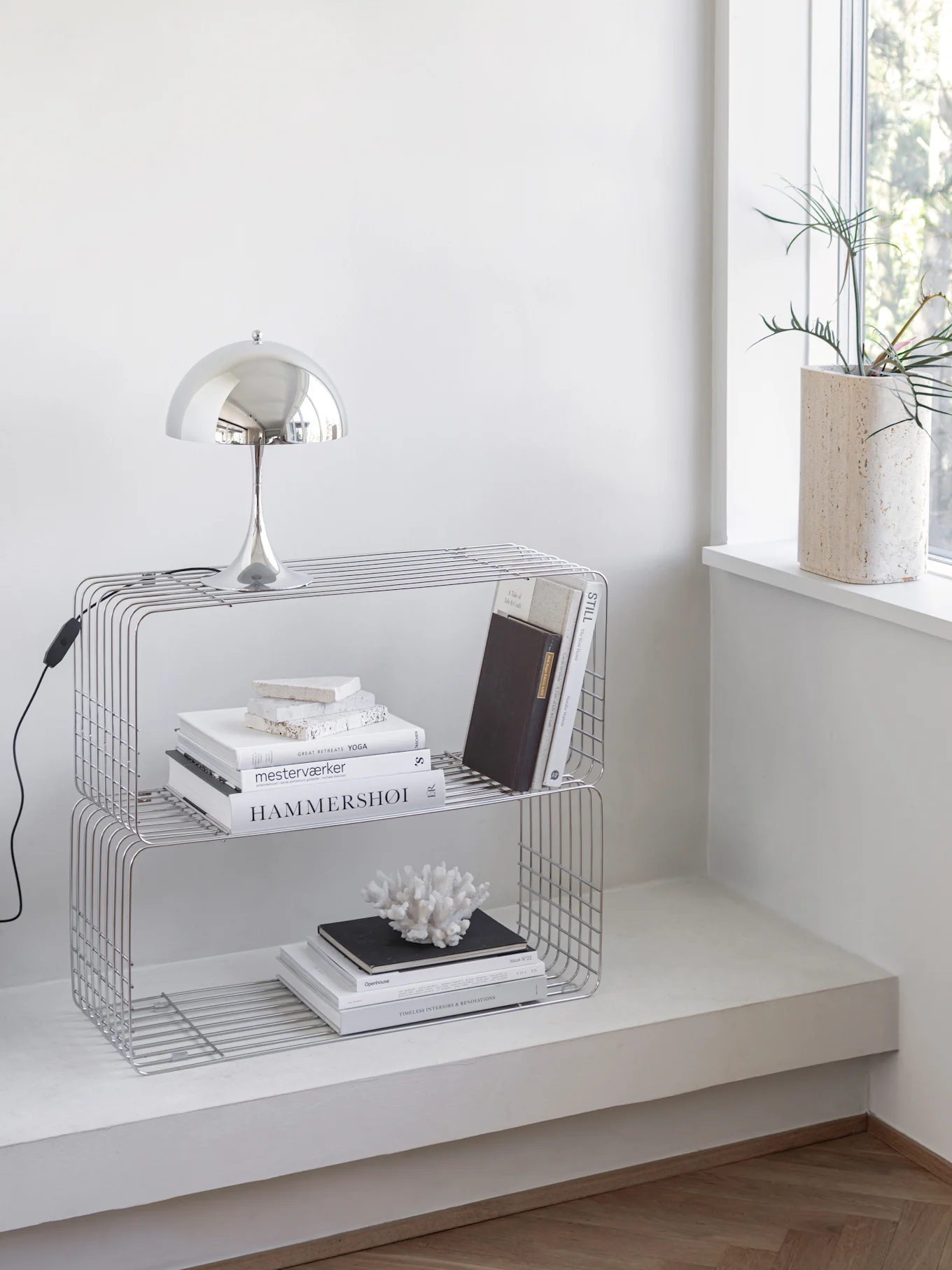 A chrome mushroom lamp sits on top of a two-tier metal wire shelf unit, which holds books and a white coral decoration. A plant in a beige pot is on the windowsill.