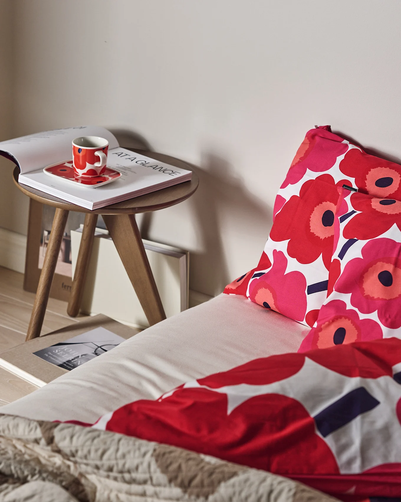 A bed with red and pink floral bedding and a textured beige throw. A red cup and open book are on a wooden side table.