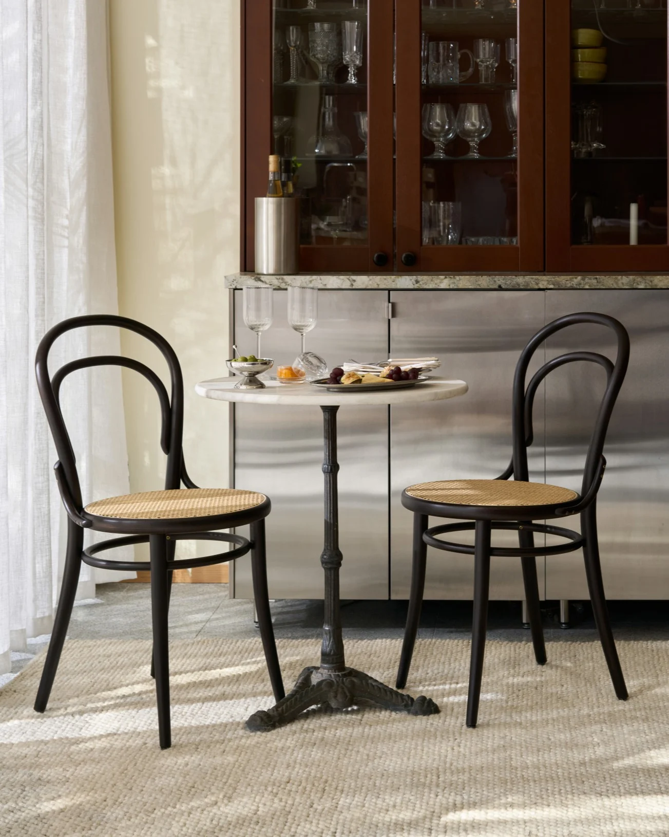 A bistro table with two dark wood and cane chairs set with snacks and wine glasses in front of a cabinet displaying glassware.